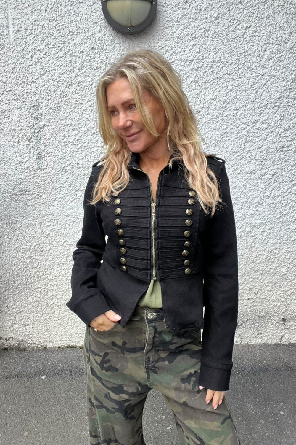 Blond kvinna står lutad mot en vit, grovputsad vägg och tittar åt sidan med ett svagt leende. Hon har svart militärinspirerad jacka med guldfärgade knappar och kamouflagebyxor; en rund vägglampa syns ovanför.