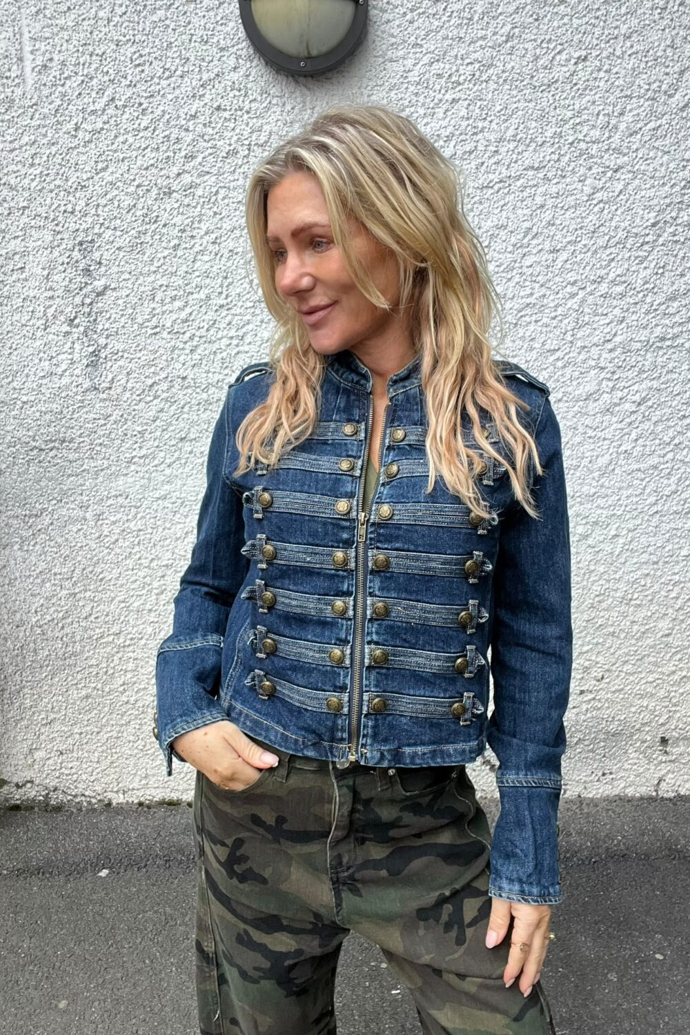 Blond kvinna står avslappnat mot en ljus, grovputsad vägg och tittar åt sidan med ett svagt leende. Hon bär en blå jeansjacka med många knappar och dragkedja samt kamouflagemönstrade byxor.
