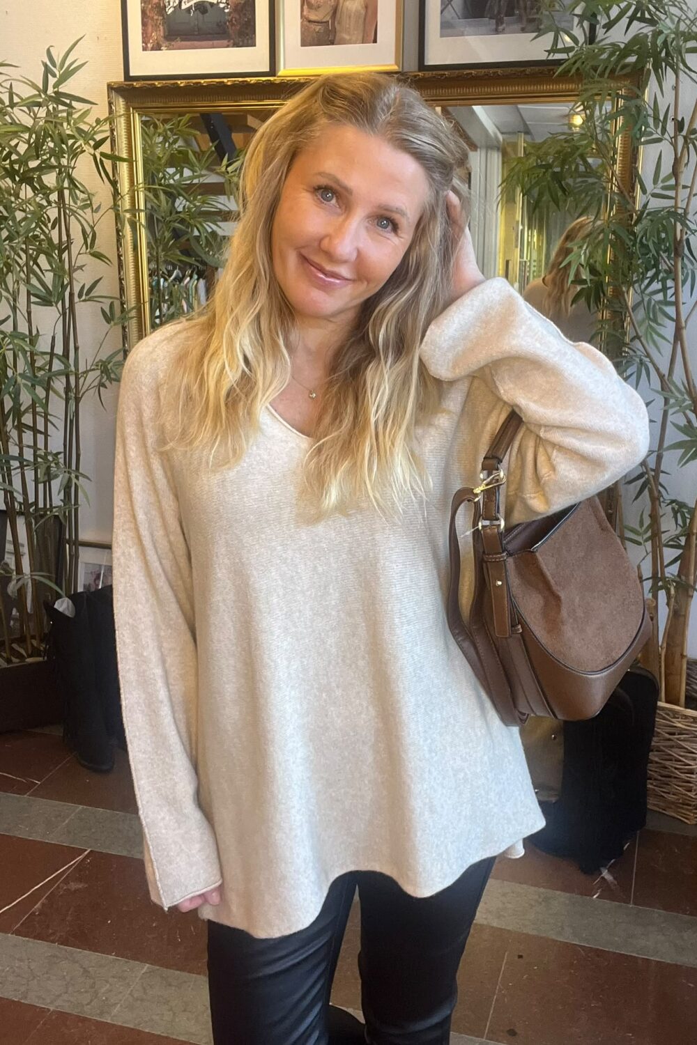 Leende kvinna med långt blont hår poserar och håller handen i håret, med en brun axelväska över armen. Hon står inomhus framför en stor guldramsspegel, omgiven av gröna växter och tavlor på väggen.