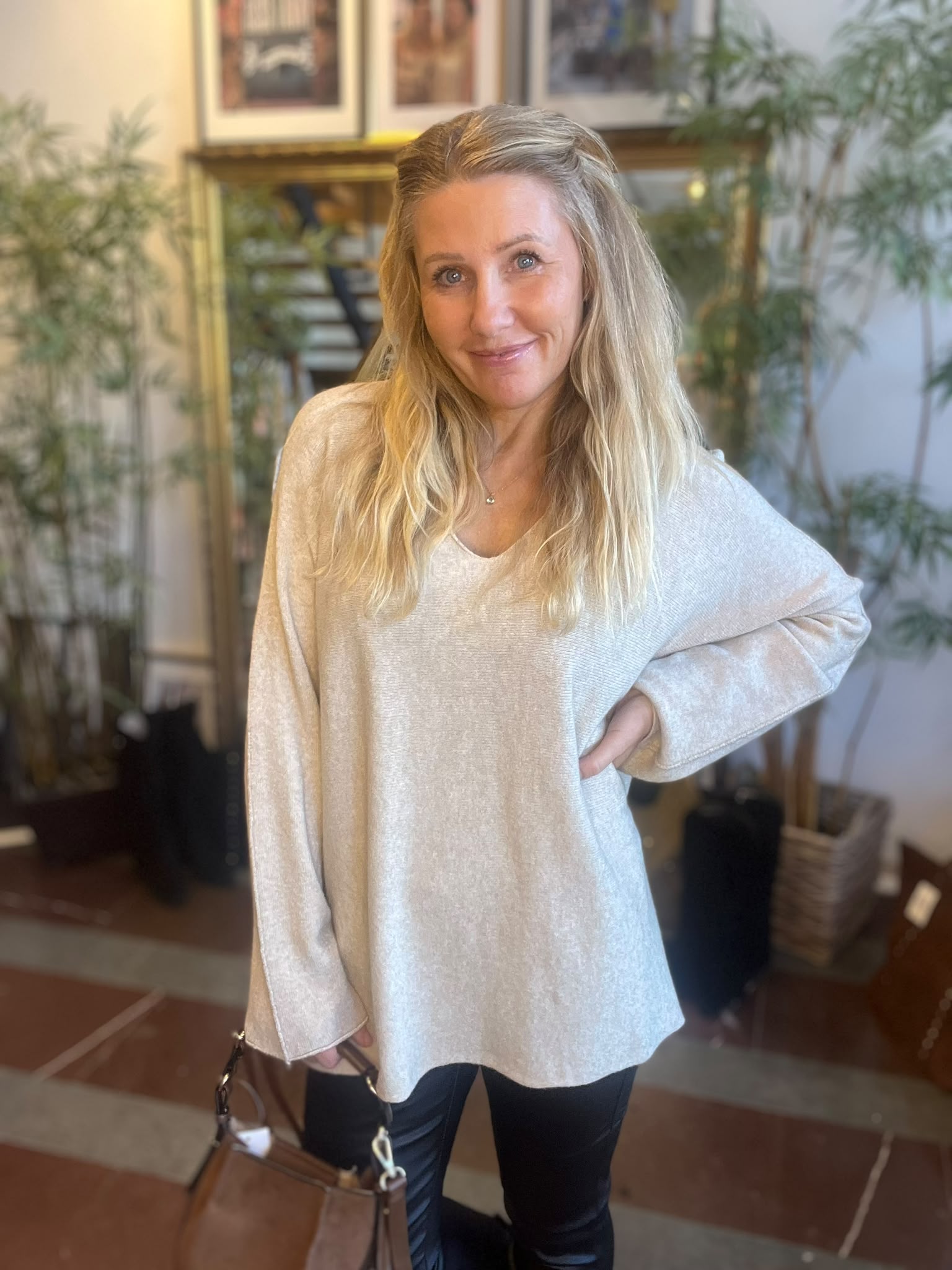 Leende kvinna med långt blont hår står inomhus med ena handen på höften och den andra vid en brun handväska. Hon bär en ljus stickad tröja och mörka byxor, med växter och tavlor i bakgrunden.