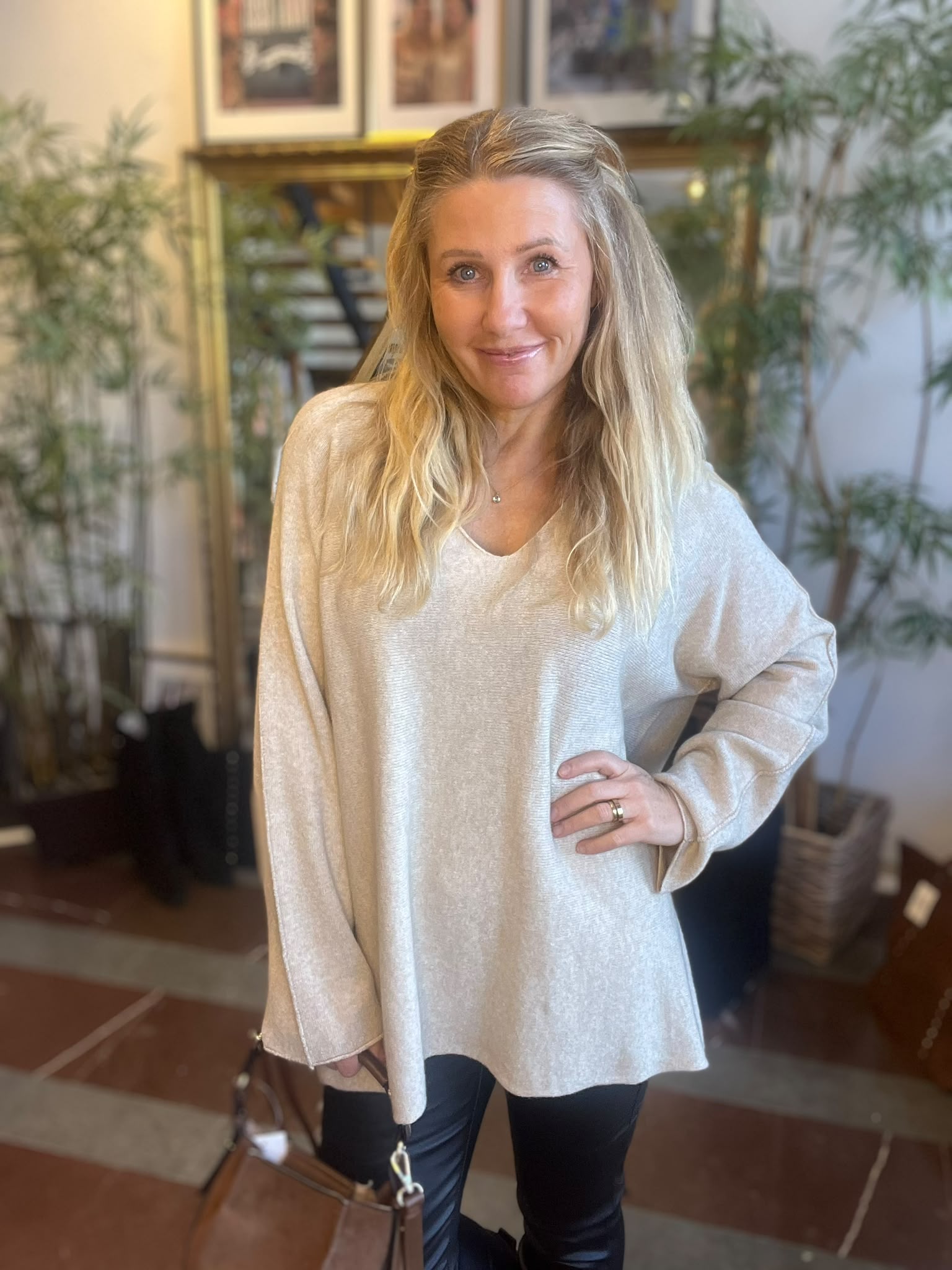 Leende kvinna med långt blont hår står inomhus med ena handen på höften och den andra vid en brun handväska. Hon bär en ljus, lös stickad tröja och svarta byxor, med växter och tavlor i bakgrunden.