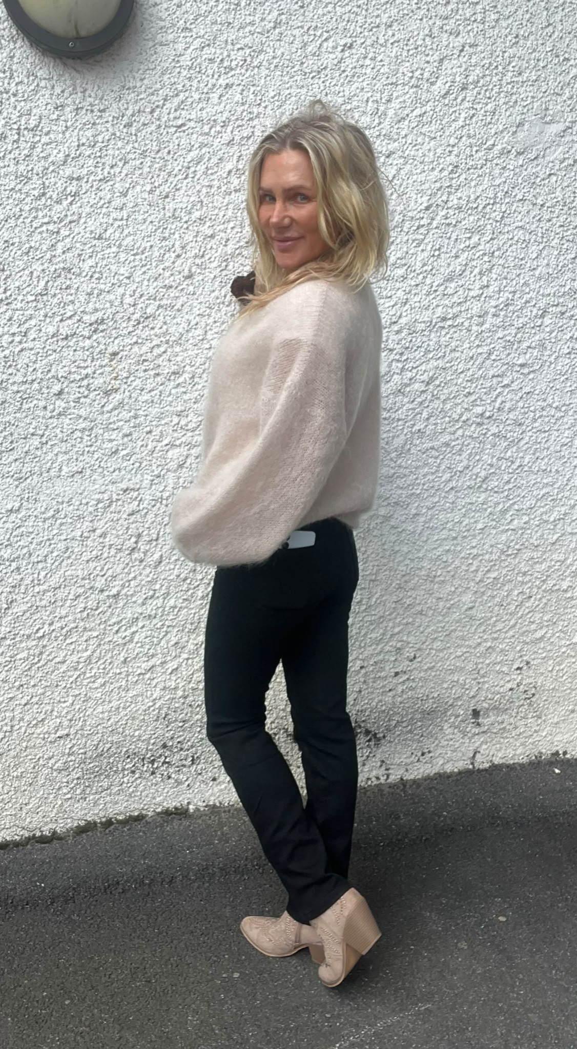 Blond kvinna står i sidoprofil och vänder sig leende mot kameran, med händerna i fickorna. Hon bär beige stickad tröja, svarta byxor och beige boots, framför en vit, grovputsad vägg utomhus.