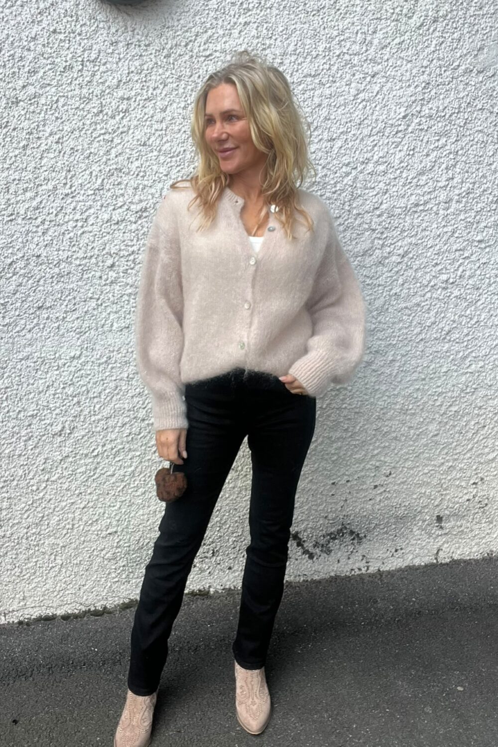 Blond kvinna står avslappnat mot en vit, grovputsad vägg och tittar åt sidan med ett leende. Hon bär ljusbeige kofta, svarta byxor och ljusa boots, och håller en liten brun accessoar i handen.
