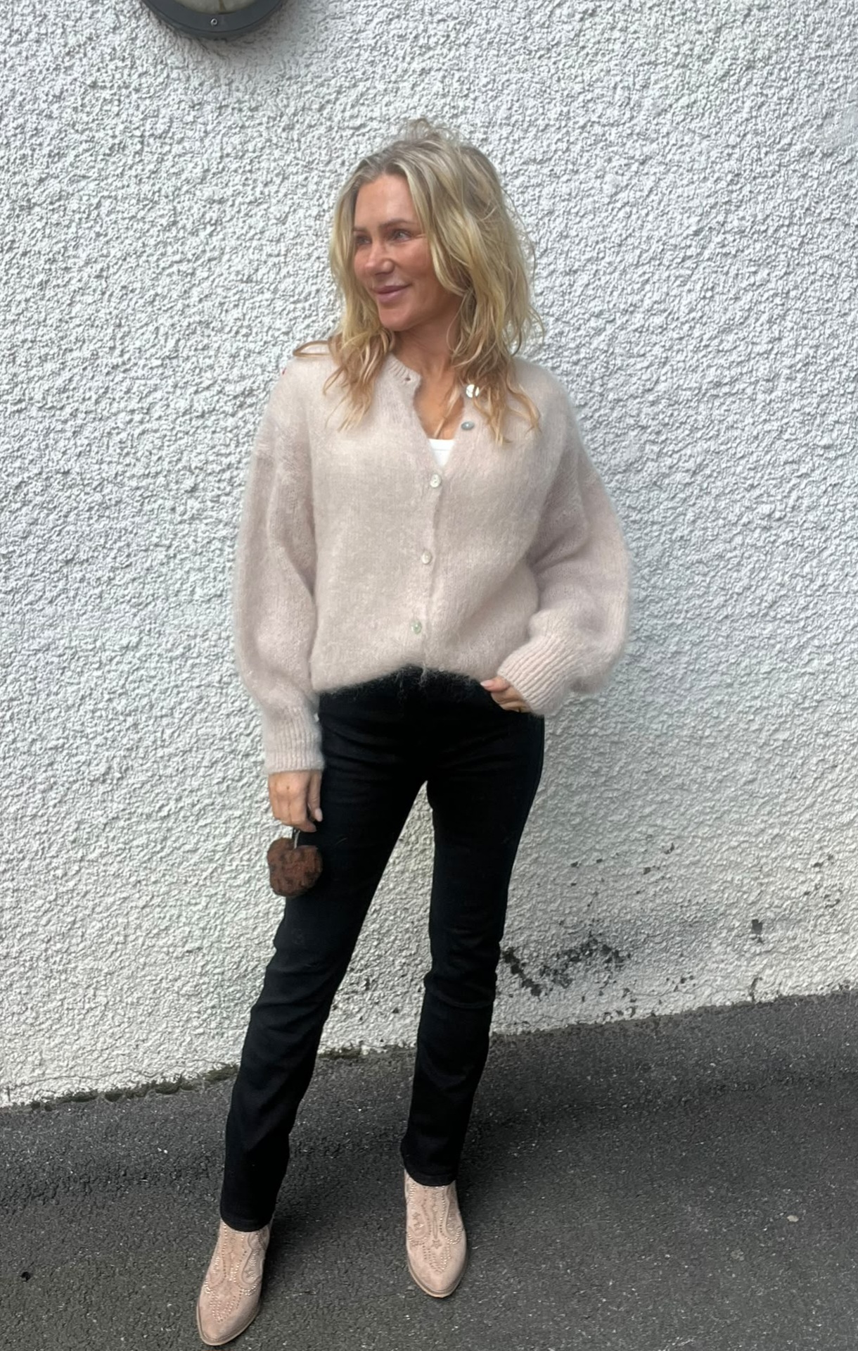 Blond kvinna står avslappnat mot en vit, grovputsad vägg och tittar åt sidan med ett leende. Hon bär ljusbeige kofta, svarta byxor och ljusa boots, och håller en liten brun accessoar i handen.