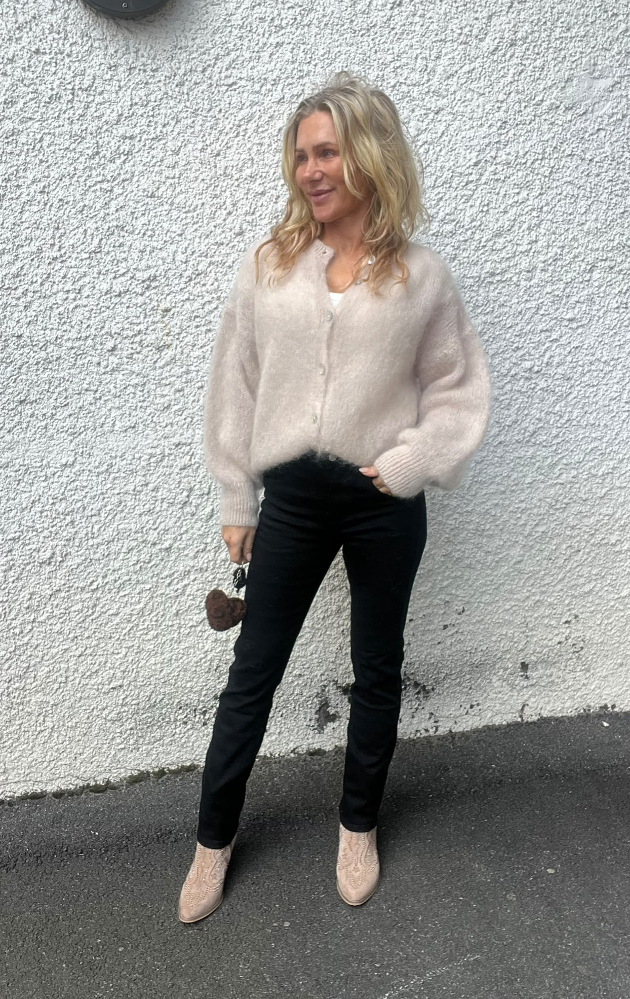 Blond kvinna står avslappnat mot en vit, grovputsad vägg och tittar åt sidan. Hon bär beige stickad kofta, svarta tighta byxor och ljusa boots, och håller en liten brun hjärtformad accessoar i handen.