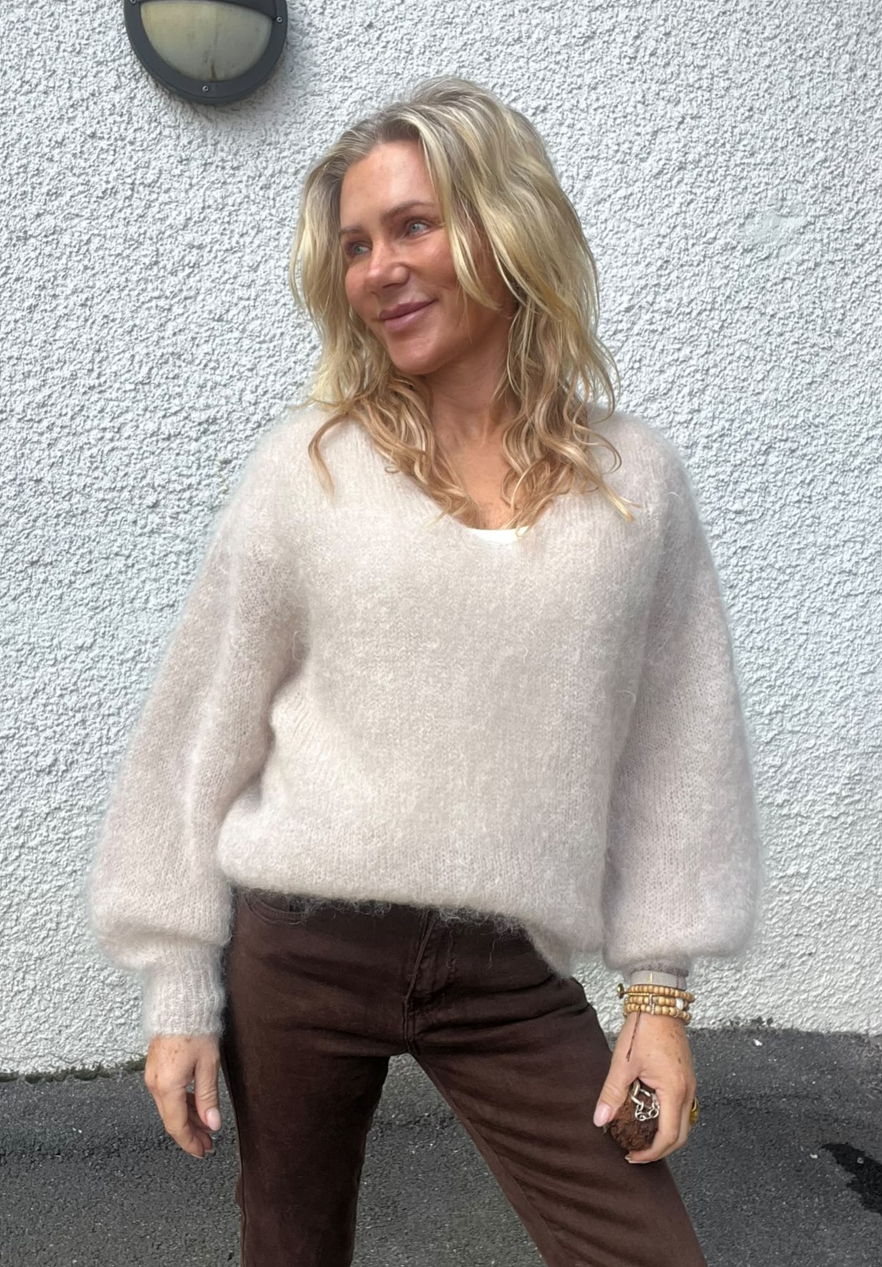 Blond kvinna står lutad mot en vit, grovputsad vägg och tittar åt sidan med ett svagt leende. Hon bär en ljus, fluffig stickad tröja och mörkbruna byxor, med armband och nycklar i handen.