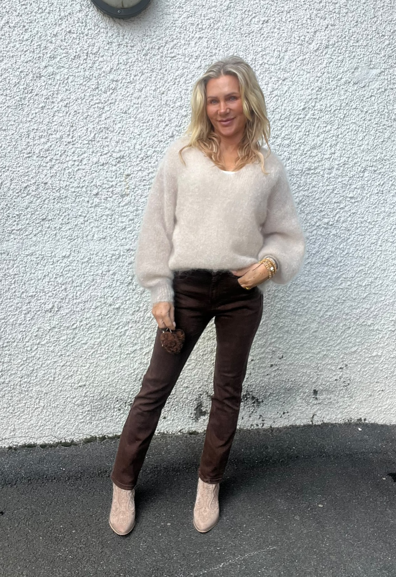 Blond kvinna står och poserar mot en vit, grovputsad vägg utomhus. Hon bär beige stickad tröja, mörkbruna byxor och ljusa boots, håller en liten brun accessoar i handen och ler mot kameran.