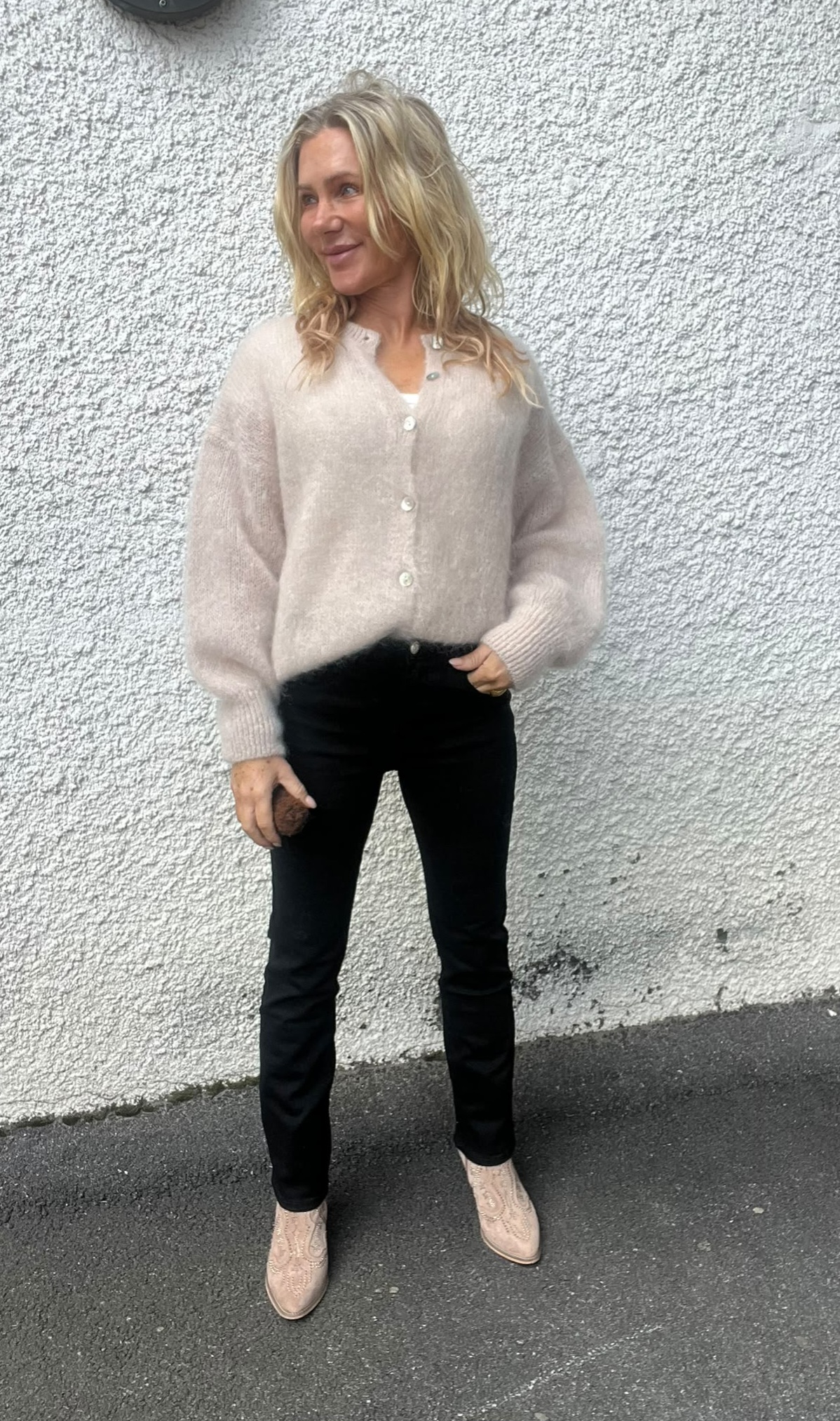 Blond kvinna står och ler med blicken åt sidan mot en ljus, grovputsad vägg. Hon har en beige stickad kofta med knappar, svarta tajta byxor och ljusa boots; ena handen i fickan.