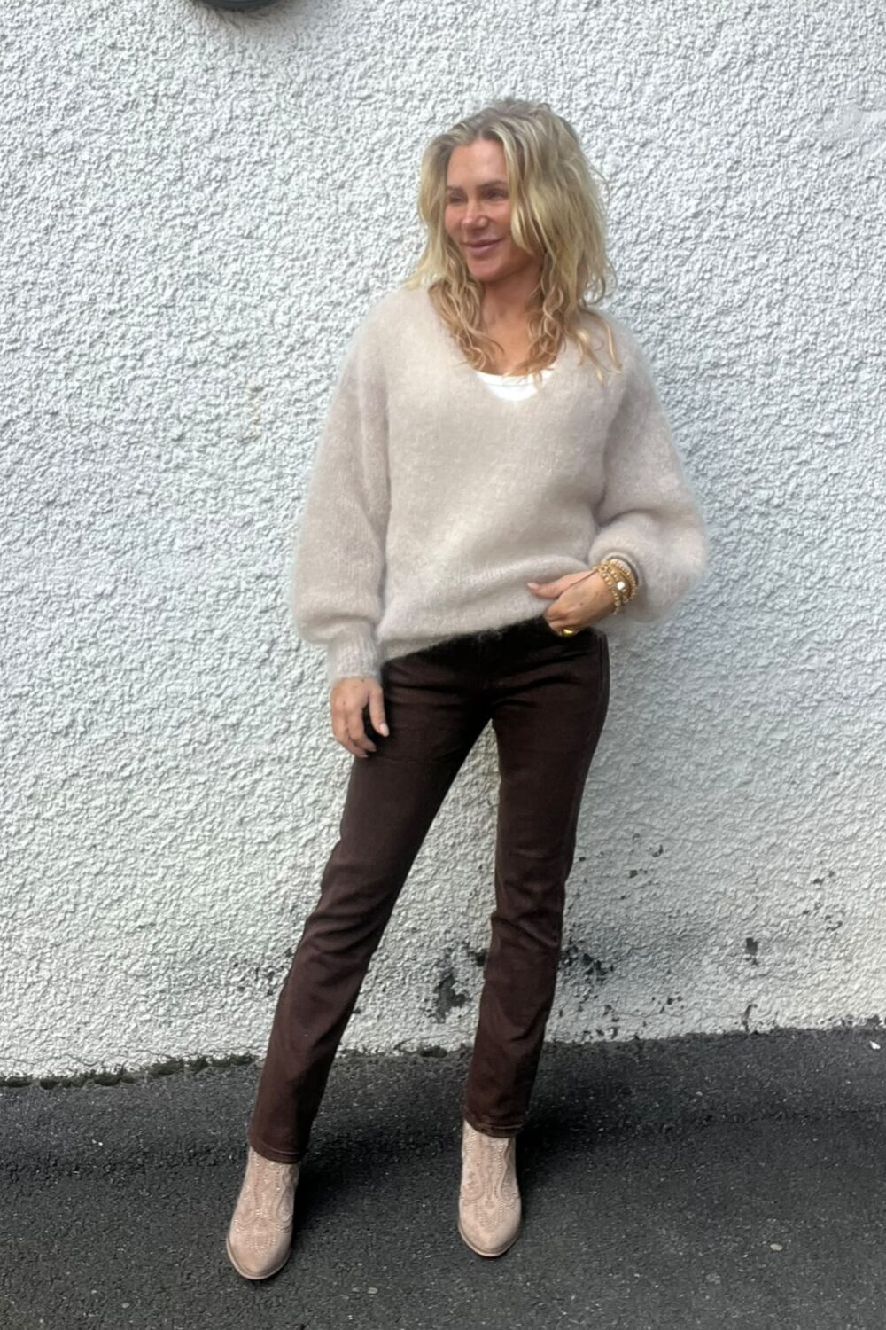 Blond kvinna står lutad mot en ljus, grovputsad vägg och tittar åt sidan. Hon bär beige stickad tröja över vit topp, mörkbruna byxor och ljusa ankelboots, med armband på handleden, på asfalt utomhus.
