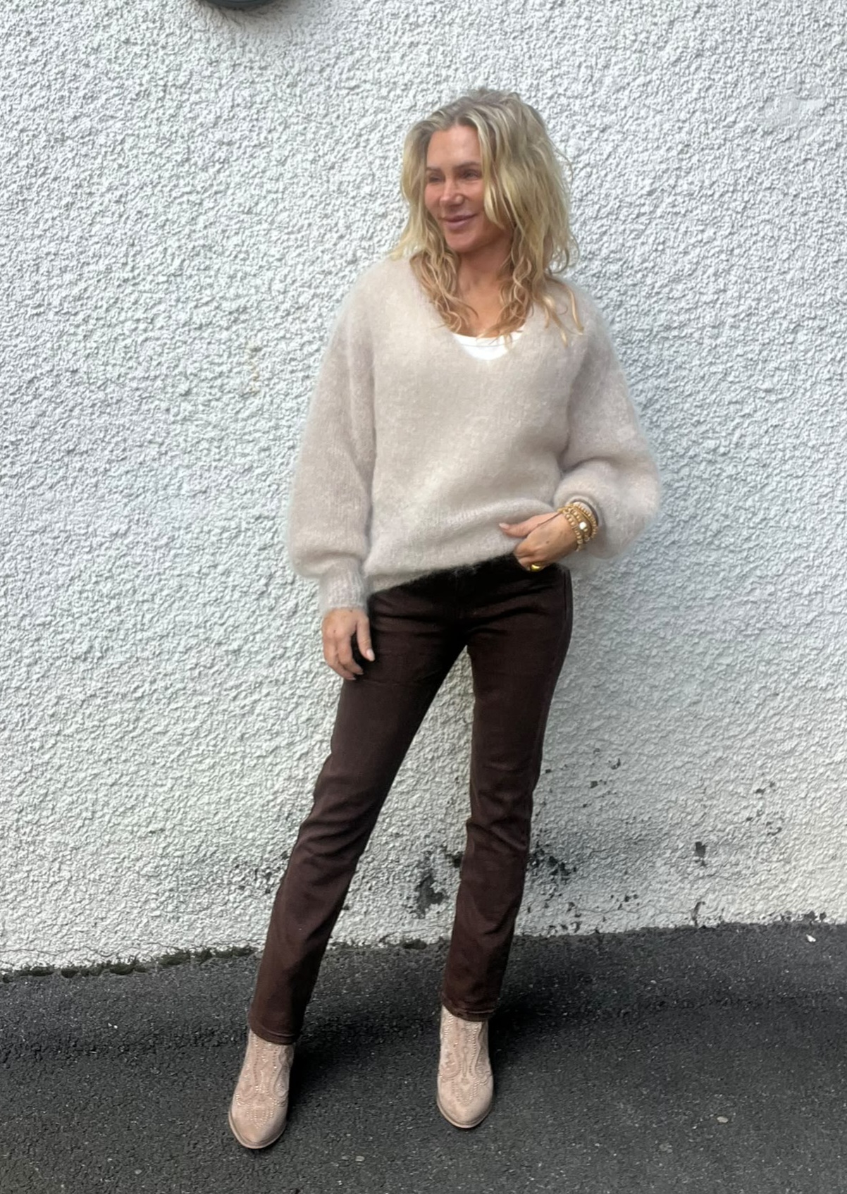 Blond kvinna står lutad mot en ljus, grovputsad vägg och tittar åt sidan. Hon bär beige stickad tröja över vit topp, mörkbruna byxor och ljusa ankelboots, med armband på handleden, på asfalt utomhus.