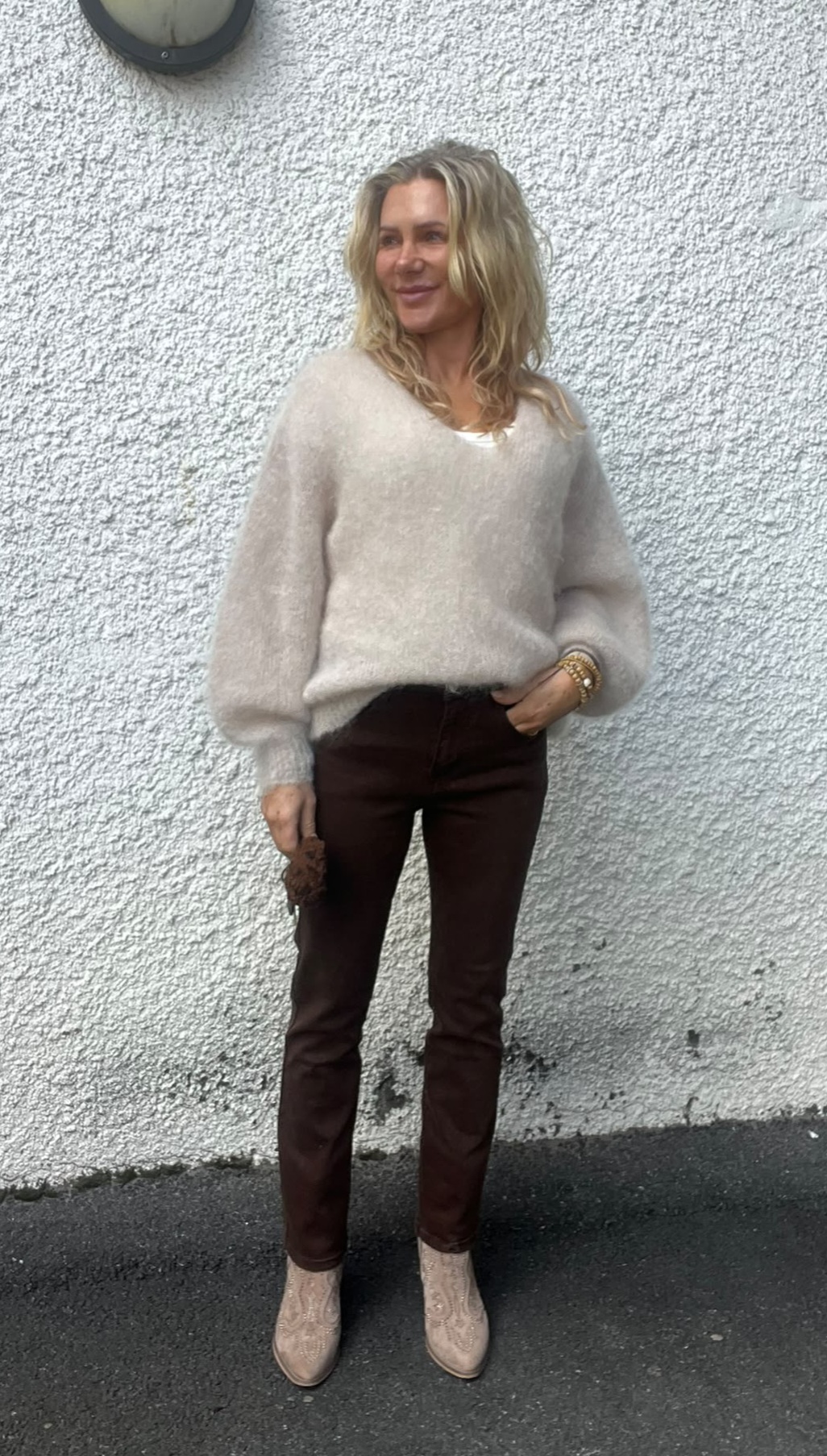 Kvinna med axellångt blont hår står och ler lätt mot sidan, lutad mot en vit, grovputsad vägg. Hon bär beige stickad tröja, bruna byxor och ljusa boots, med en hand i fickan.