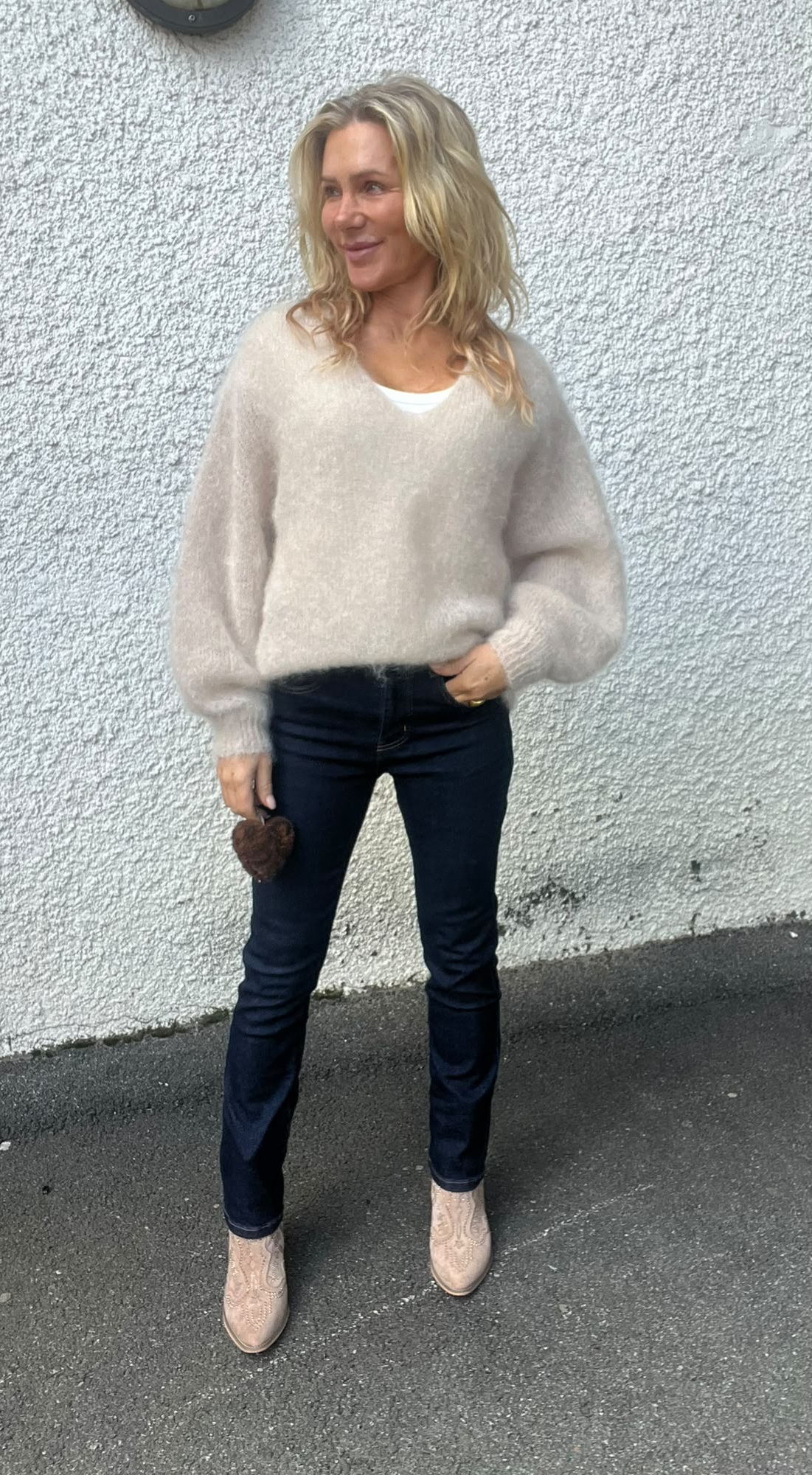 Blond kvinna står avslappnat och ler medan hon tittar åt sidan, framför en vit, grovputsad vägg. Hon bär beige stickad tröja, mörka jeans och ljusa boots, och håller en brun pälsboll i handen.