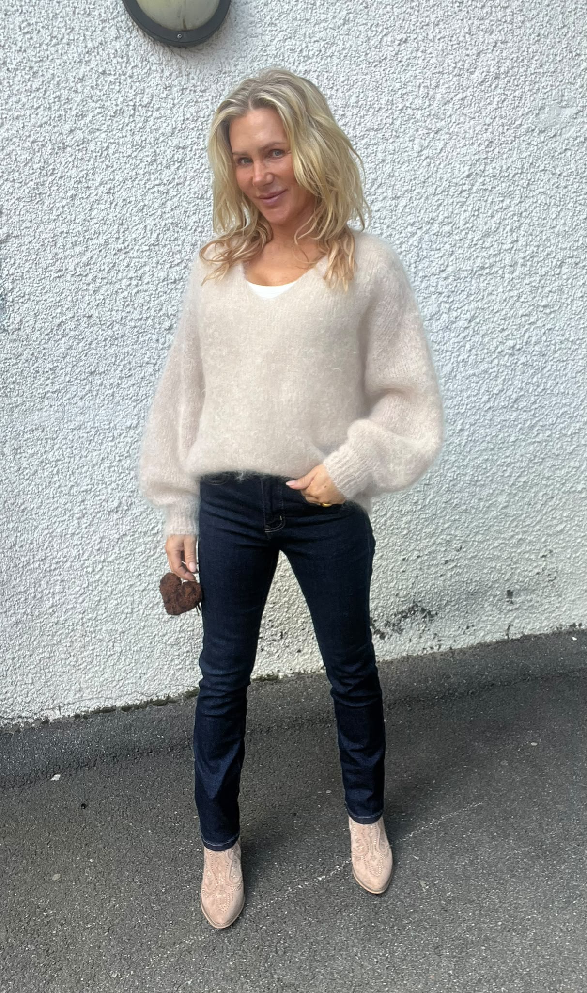 Blond kvinna står och poserar mot en vit, grovputsad vägg utomhus, med ena handen i jeansfickan och den andra hållande en liten brun accessoar. Hon bär beige stickad tröja, mörka jeans och ljusa boots.