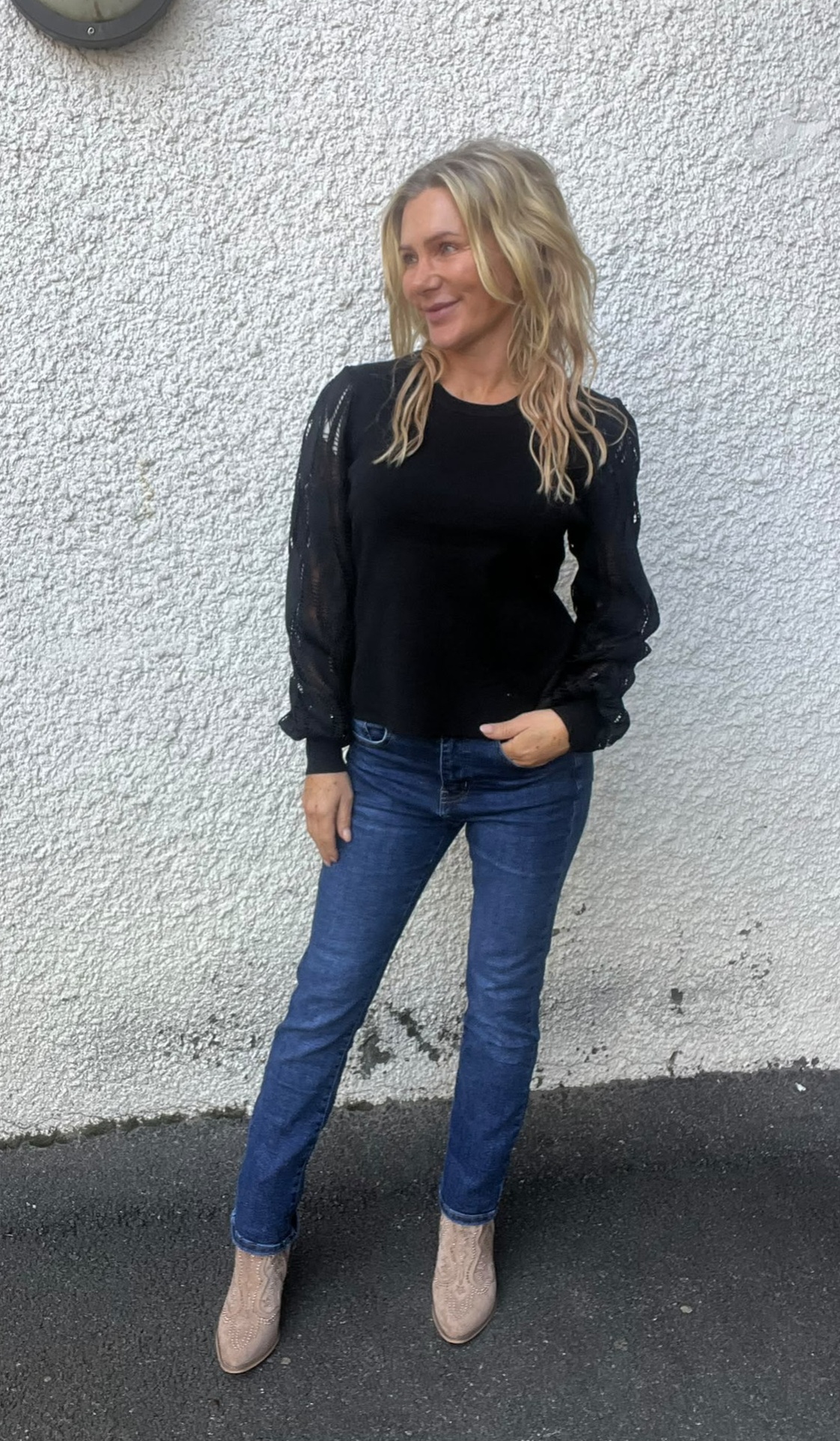 Blond kvinna står avslappnat mot en vit, grovputsad vägg och ler åt sidan, med en hand i jeansfickan. Hon bär svart topp med genomskinliga ärmar, blå jeans och beige boots på asfalt utomhus.