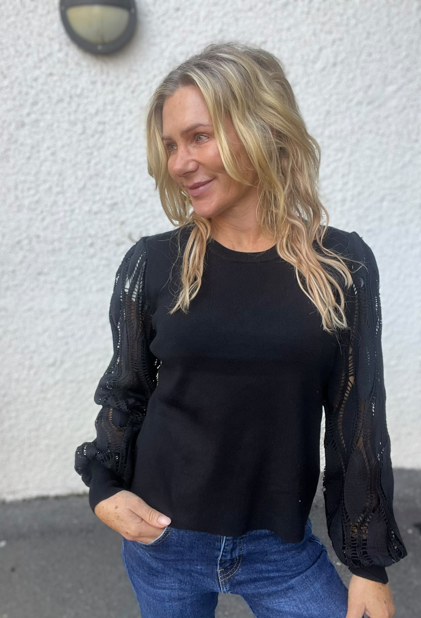 Leende kvinna med blont, vågigt hår står och tittar åt sidan, med ena handen i jeansfickan. Hon bär en svart topp med genombrutna spetsärmar och står framför en vit, grovputsad vägg.
