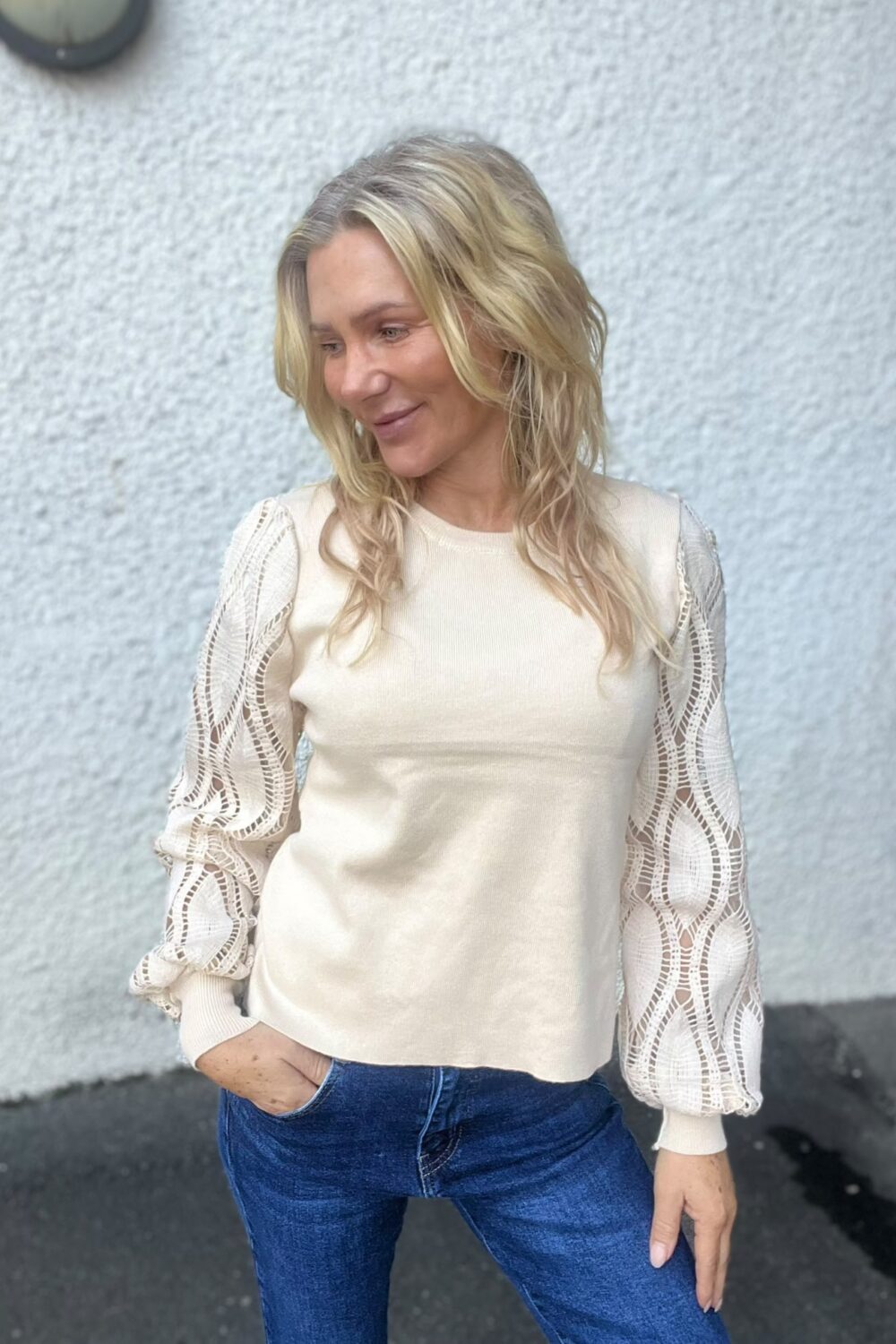 Blond kvinna står lutad mot en ljus, grovputsad vägg och tittar ned åt sidan med ett svagt leende. Hon har en beige stickad tröja med spetsiga, puffiga ärmar och blå jeans, ena handen i fickan.