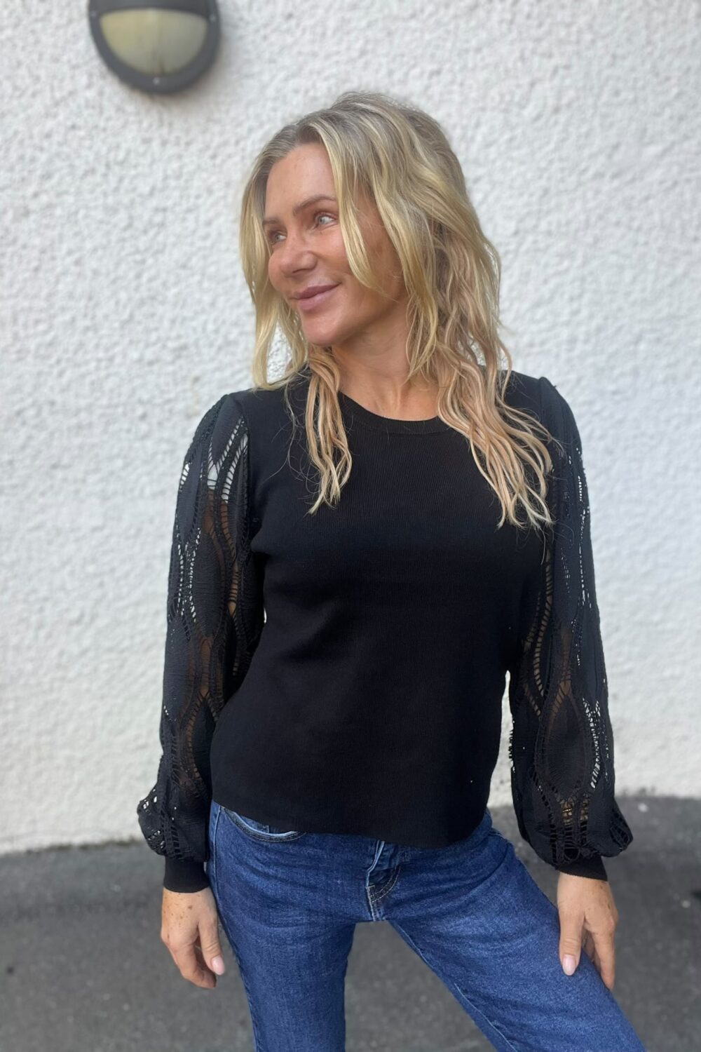 Leende kvinna med långt blont hår står och tittar åt vänster, klädd i svart topp med genombrutna spetsärmar och blå jeans. Hon poserar utomhus framför en vit putsad vägg med en vägglampa ovanför.