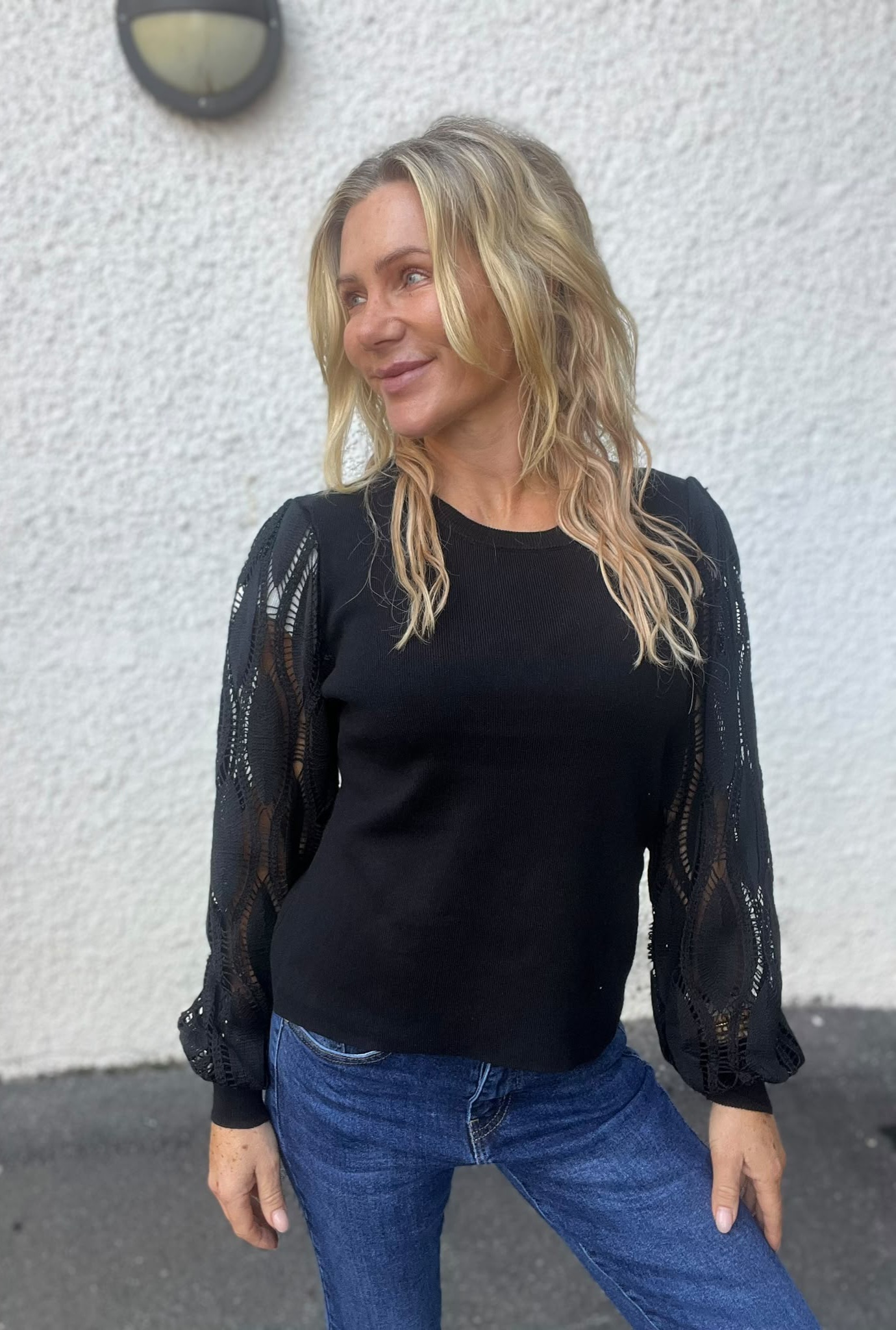 Kvinna med långt blont hår står utomhus framför en vit putsad vägg och ler medan hon tittar åt sidan. Hon bär svart stickad tröja med genombrutna ärmar och blå jeans; en vägglampa syns ovanför.
