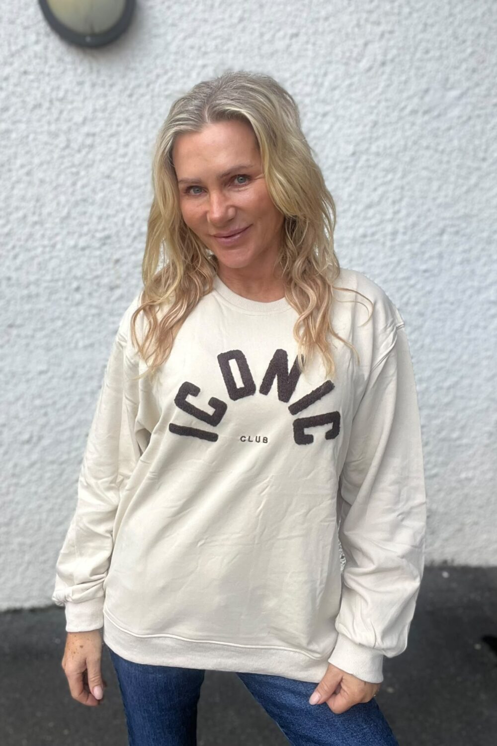 Leende blond kvinna står lätt lutad mot kameran framför en ljus, grovputsad vägg utomhus. Hon bär en ljus sweatshirt med texten “ICONIC CLUB” och jeans, med händerna avslappnat vid sidorna.
