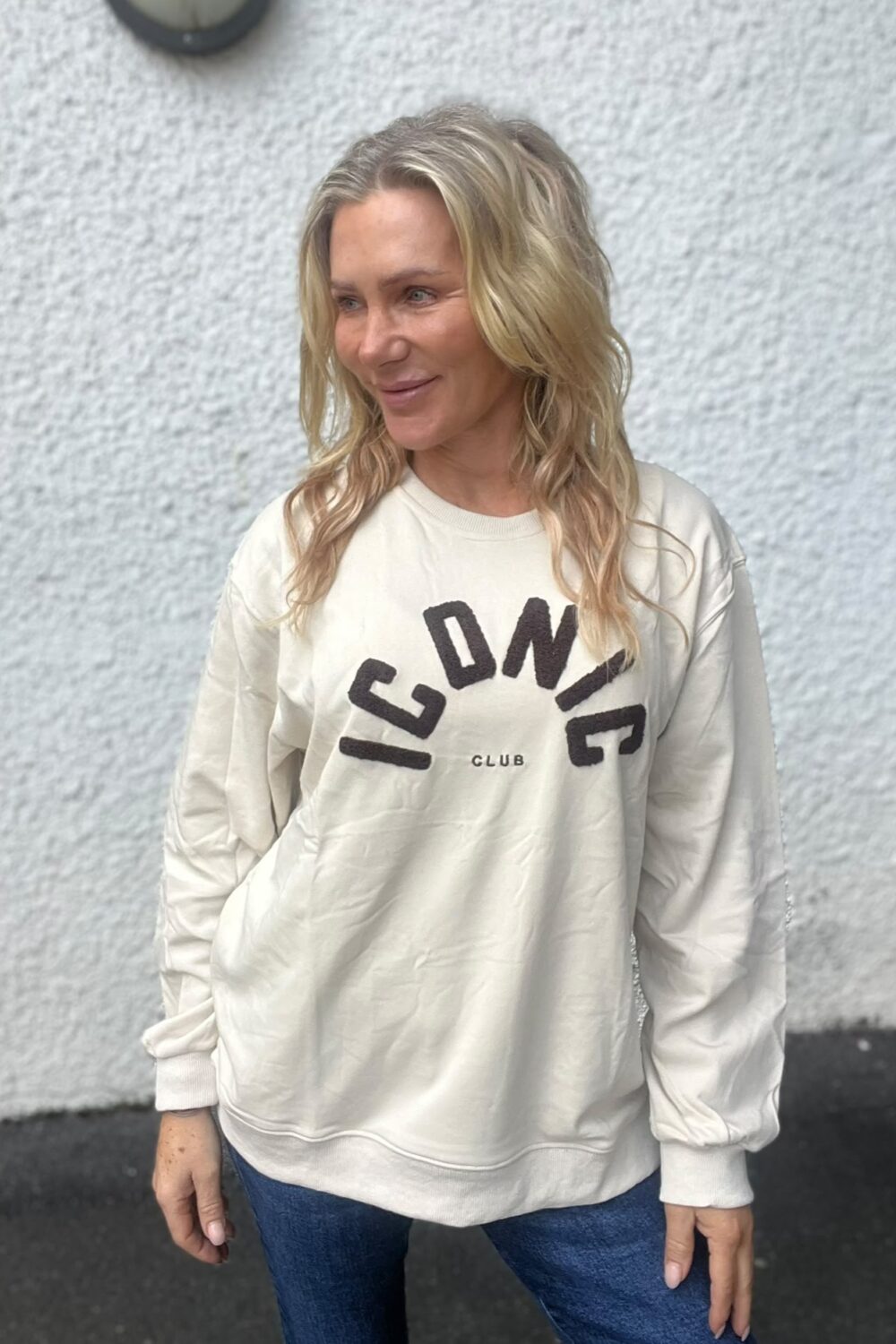 Kvinna med långt blont hår står och ler lätt medan hon tittar åt sidan, mot en ljus, grovputsad vägg. Hon bär en vit sweatshirt med texten “ICONIC” och “CLUB” samt blå jeans.