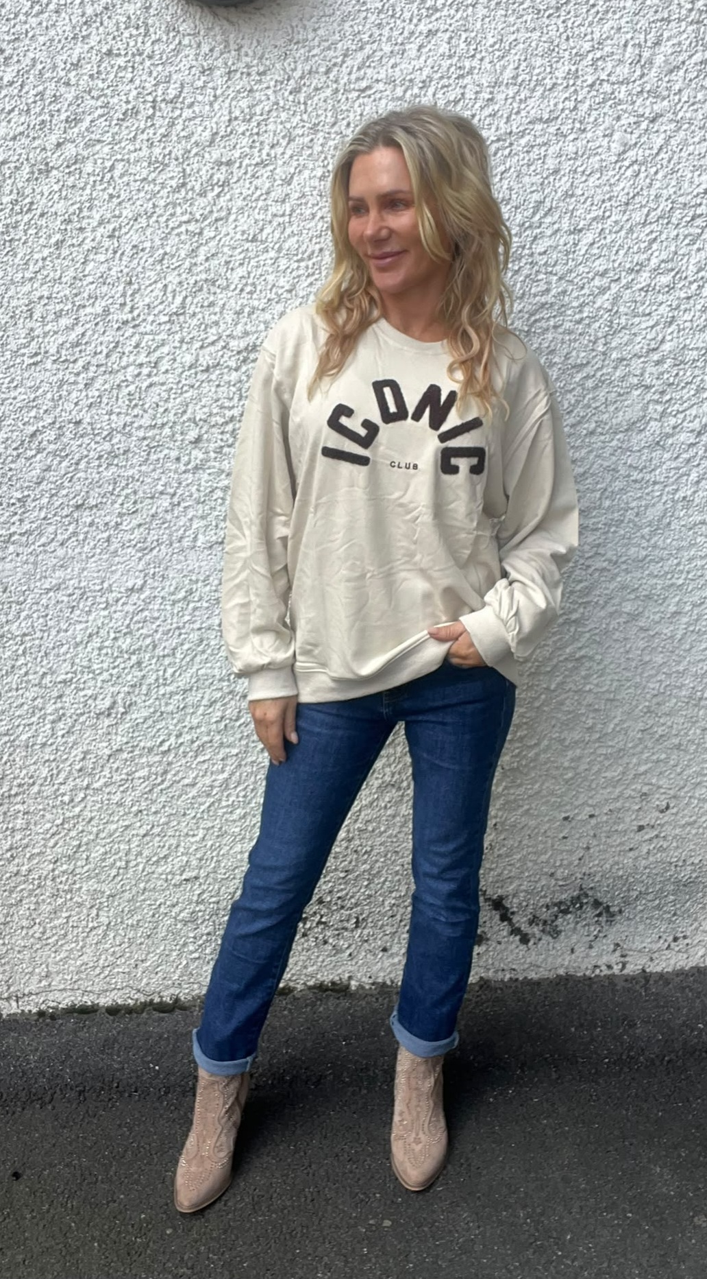Kvinna med långt blont hår står avslappnat mot en vit, grovputsad vägg och tittar åt sidan. Hon bär beige tröja med texten ”ICONIC CLUB”, blå jeans och ljusa boots, ena handen i fickan.