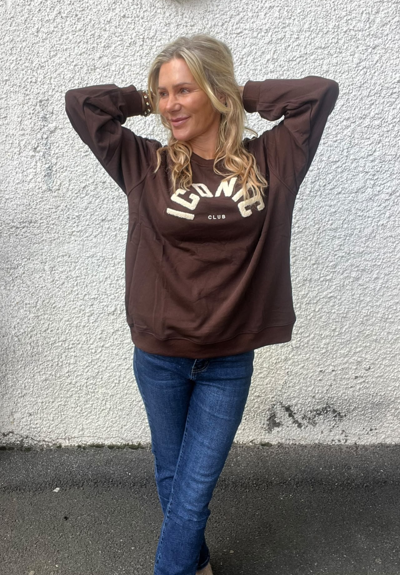 Blond kvinna står avslappnat med händerna bakom huvudet och ler åt sidan, klädd i brun sweatshirt med texten ”ICOWS CLUB” och blå jeans, framför en ljus, grovputsad vägg utomhus.