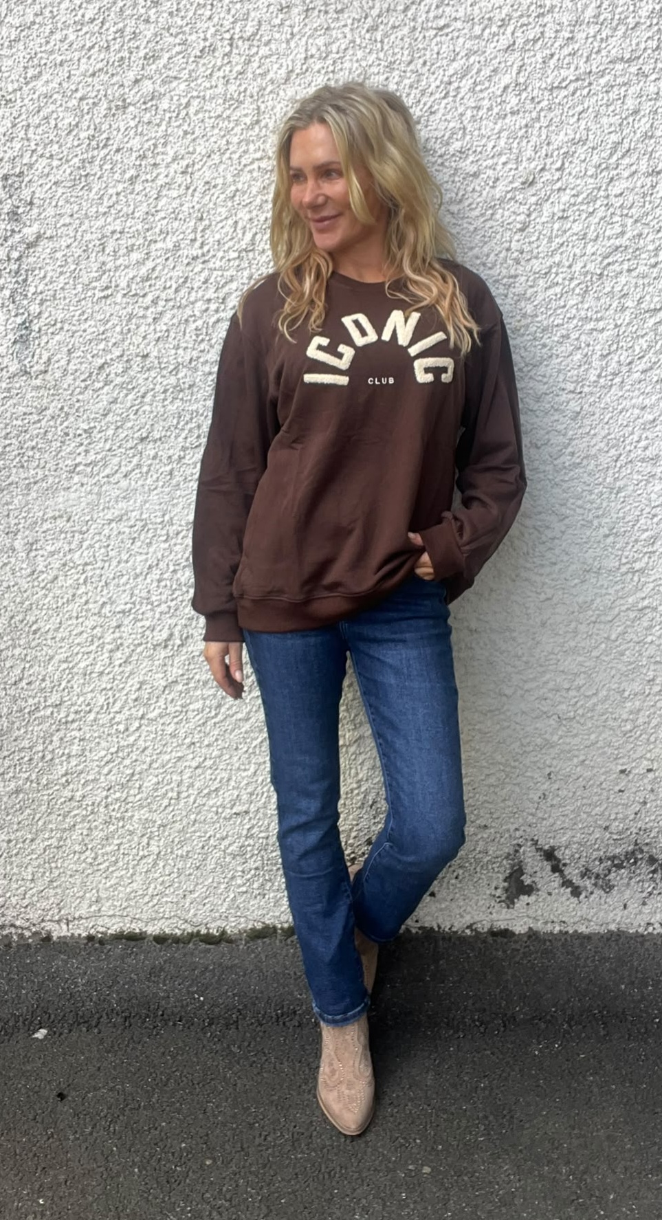 Kvinna med långt blont hår står lutad mot en vit, grov putsad vägg och tittar åt sidan. Hon bär brun sweatshirt med texten “ICONIC CLUB”, blå jeans och beige boots, en hand i fickan.