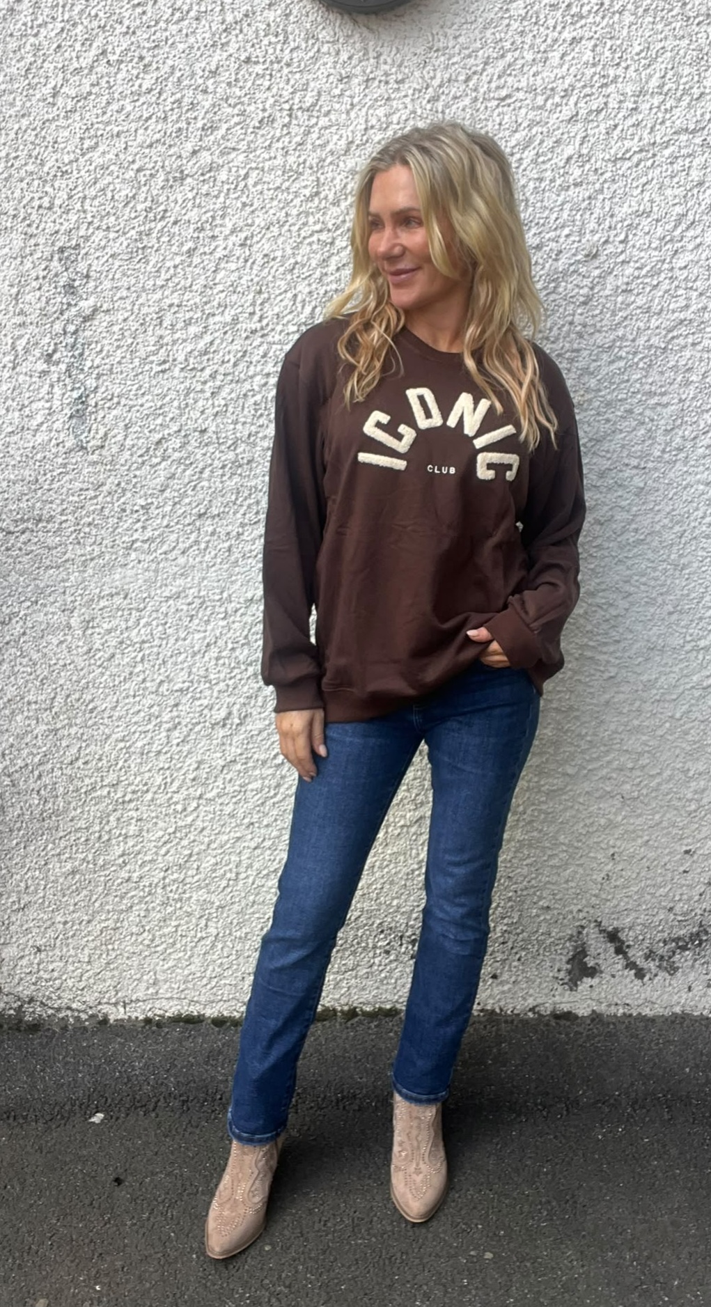 Kvinna med långt blont hår står avslappnat mot en ljus, grovputsad vägg och tittar åt sidan. Hon bär brun sweatshirt med texten ”ICONIC CLUB”, blå jeans och ljusa boots på asfalt utomhus.