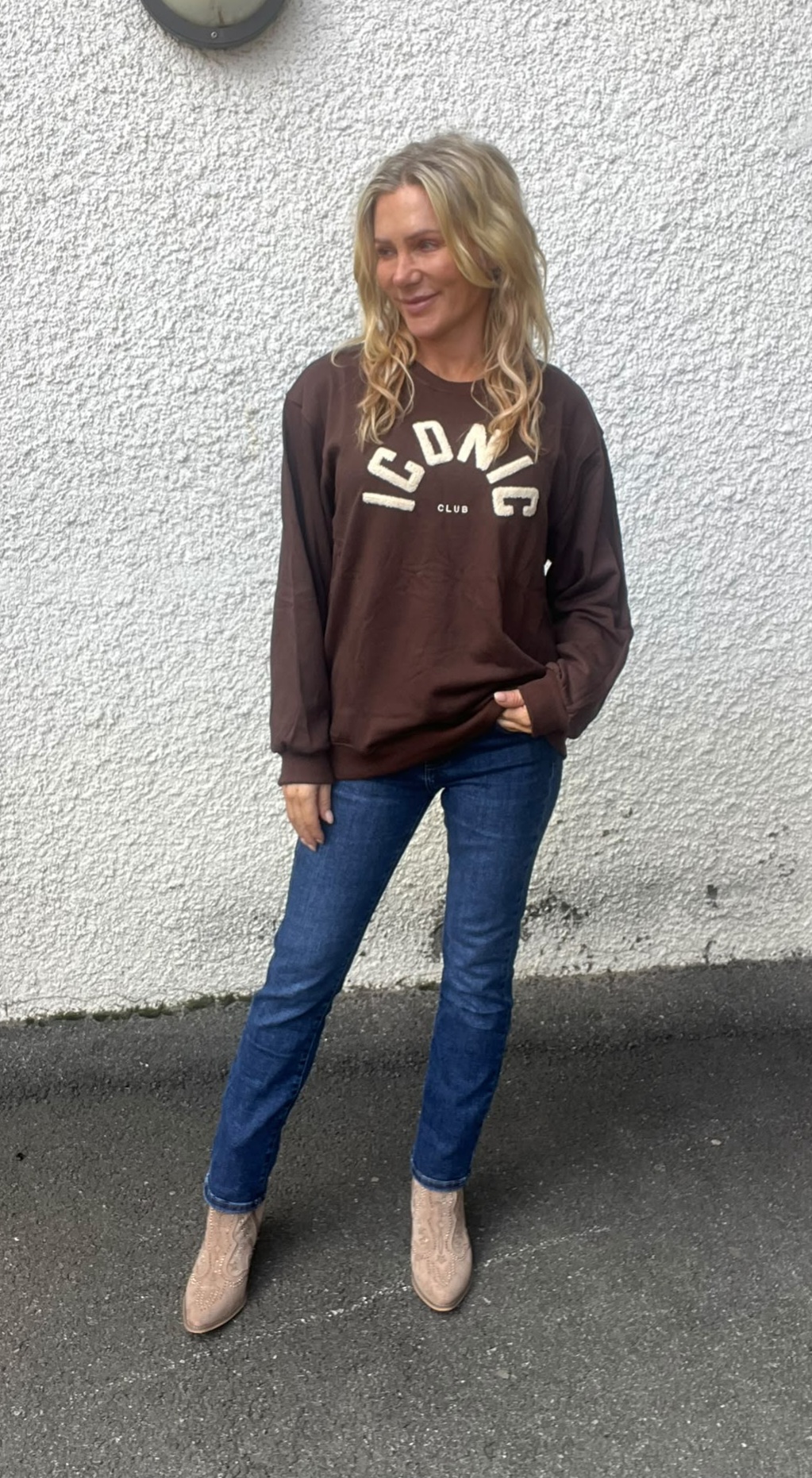 Kvinna står poserande mot en ljus, grovputsad vägg, med ena handen i fickan och blicken åt sidan. Hon bär brun sweatshirt med texten “ICONIC CLUB”, blå jeans och beige boots på asfalt.