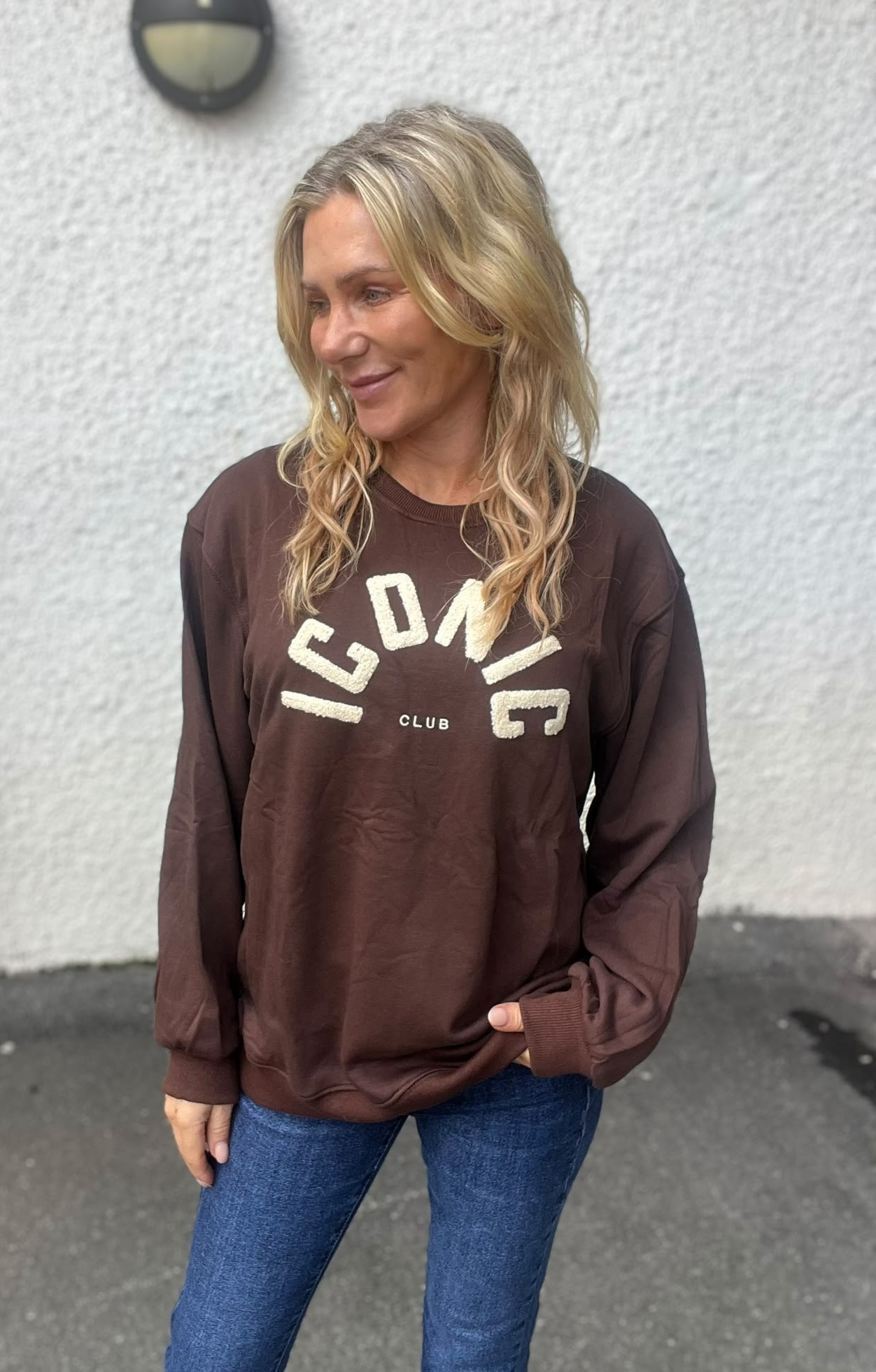 Blond kvinna står utomhus och ler medan hon tittar åt sidan, med ena handen i fickan. Hon bär brun sweatshirt med texten ”ICONIC CLUB” och blå jeans, framför en ljus, grov vägg.