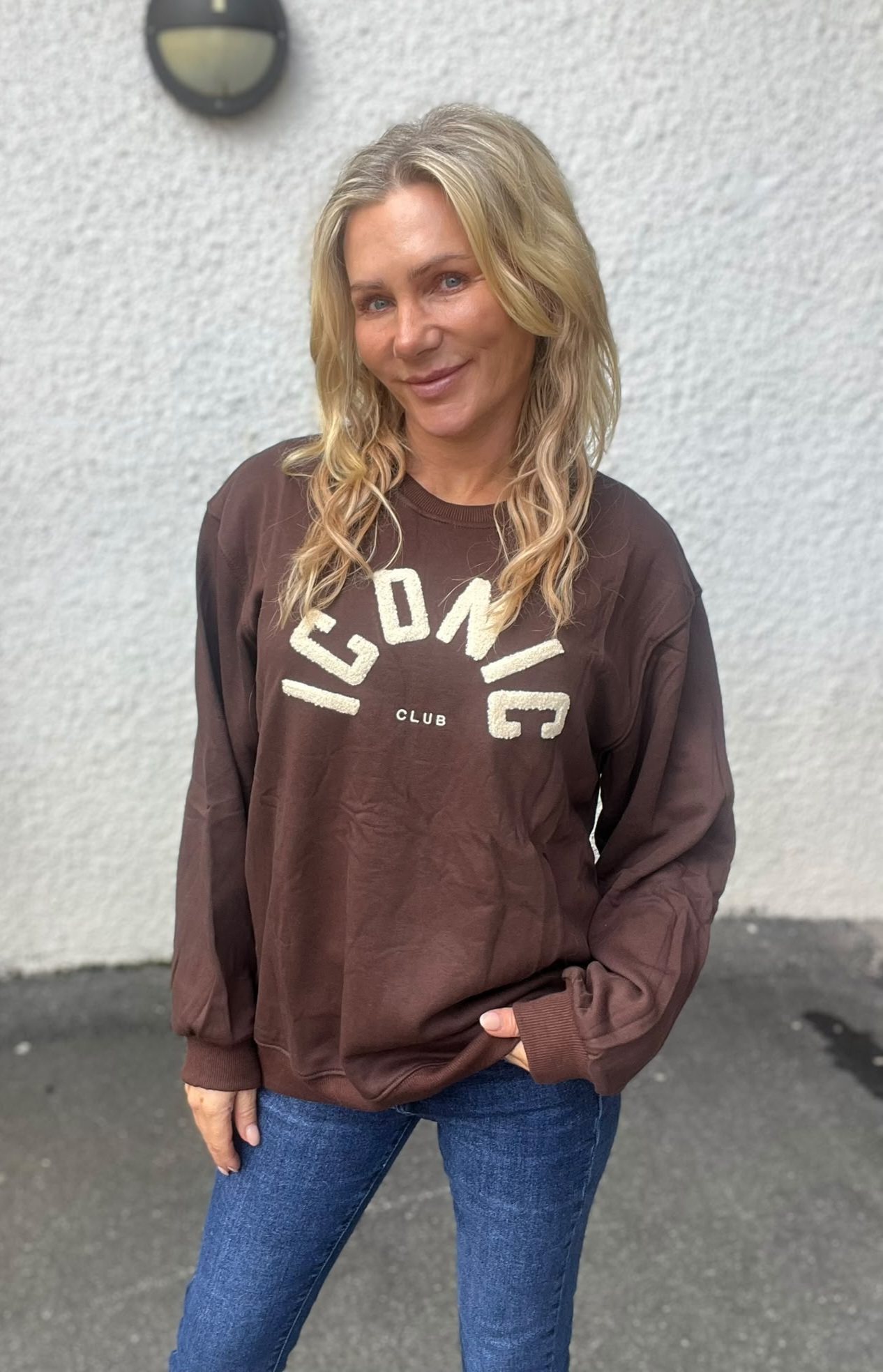 Leende kvinna med långt blont hår poserar utomhus mot en ljus vägg, med ena handen i fickan. Hon bär brun sweatshirt med texten ”ICONIC CLUB” och blå jeans; en vägglampa syns ovanför.