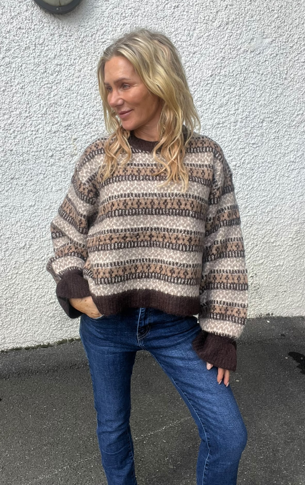 Blond kvinna står avslappnat med ena handen i jeansfickan och ler åt sidan. Hon bär en stickad tröja med bruna och beige mönsterränder. Bakgrunden är en vit putsad vägg utomhus.