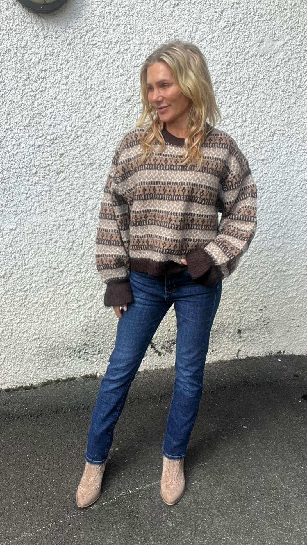 Blond kvinna står avslappnat utomhus mot en ljus, grovputsad vägg och tittar åt sidan. Hon bär mönstrad stickad tröja i bruna och beige toner, blå jeans och ljusa boots. Inga synliga texter.