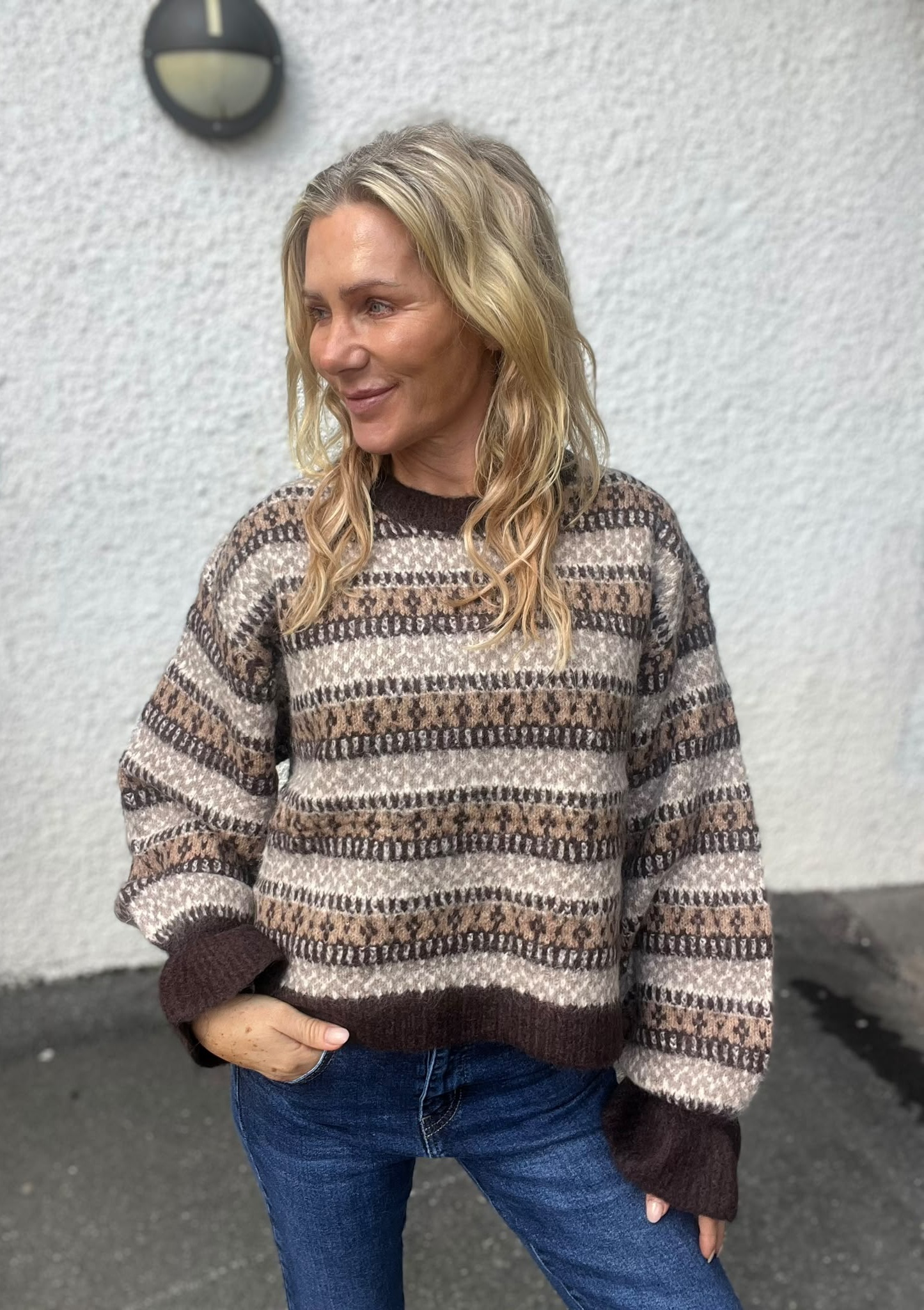 Blond kvinna står utomhus och tittar åt sidan med ett svagt leende. Hon bär en mönstrad stickad tröja i bruna och beige toner samt jeans, framför en vit putsad vägg med en rund vägglampa.