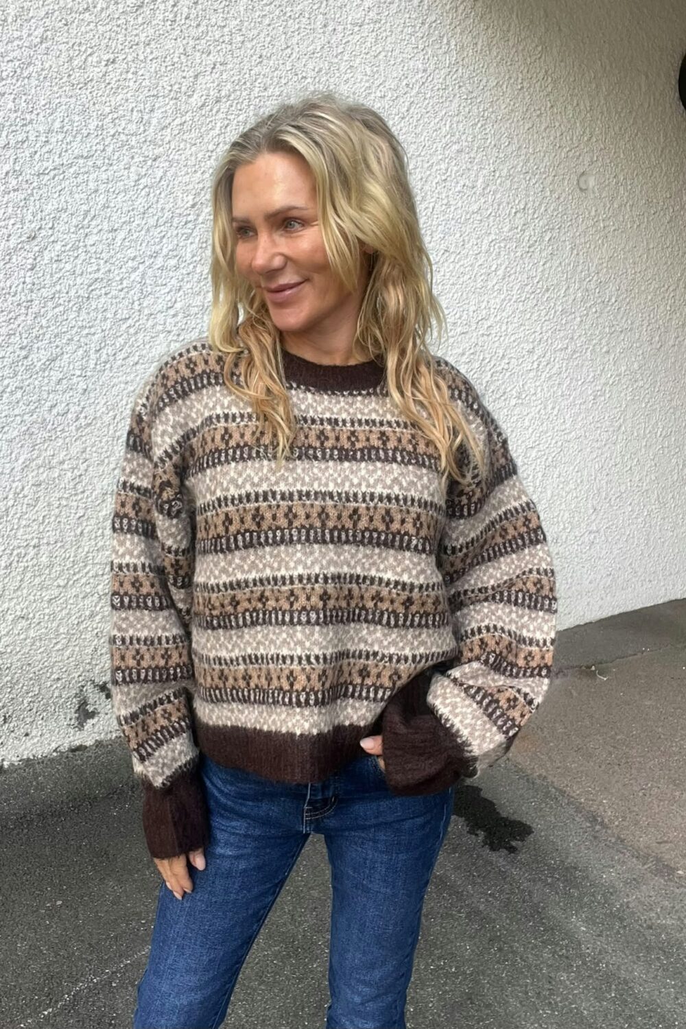 Blond kvinna står utomhus mot en vit, grovputsad vägg och ler medan hon tittar åt sidan. Hon bär en mönstrad stickad tröja i brunt och beige samt blå jeans, med en hand i fickan.