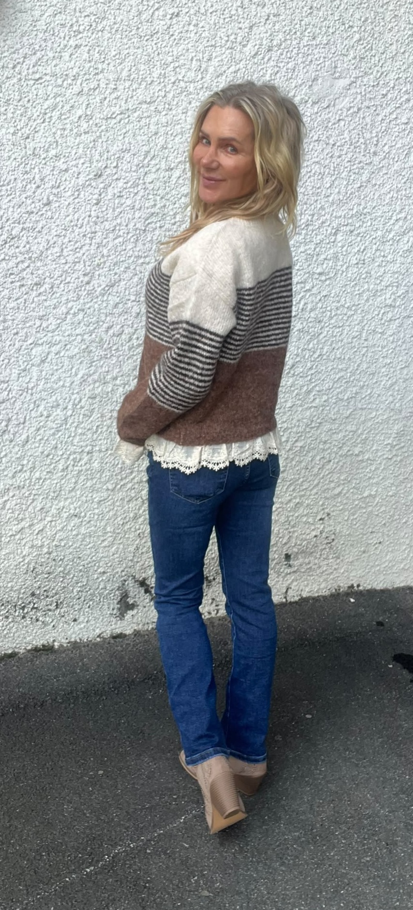 Kvinna står med ryggen delvis vänd och tittar leende över axeln mot kameran, framför en vit, grovputsad vägg. Hon bär randig stickad tröja med spetskant, blå jeans och beige klackskor på asfalt.