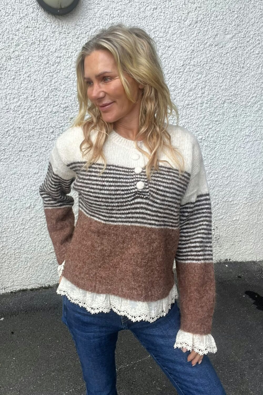 Blond kvinna står lutad mot en vit, grovputsad vägg och tittar åt vänster med ett svagt leende. Hon bär en stickad tröja med vita, svarta och bruna ränder, spetskantad blus och jeans.