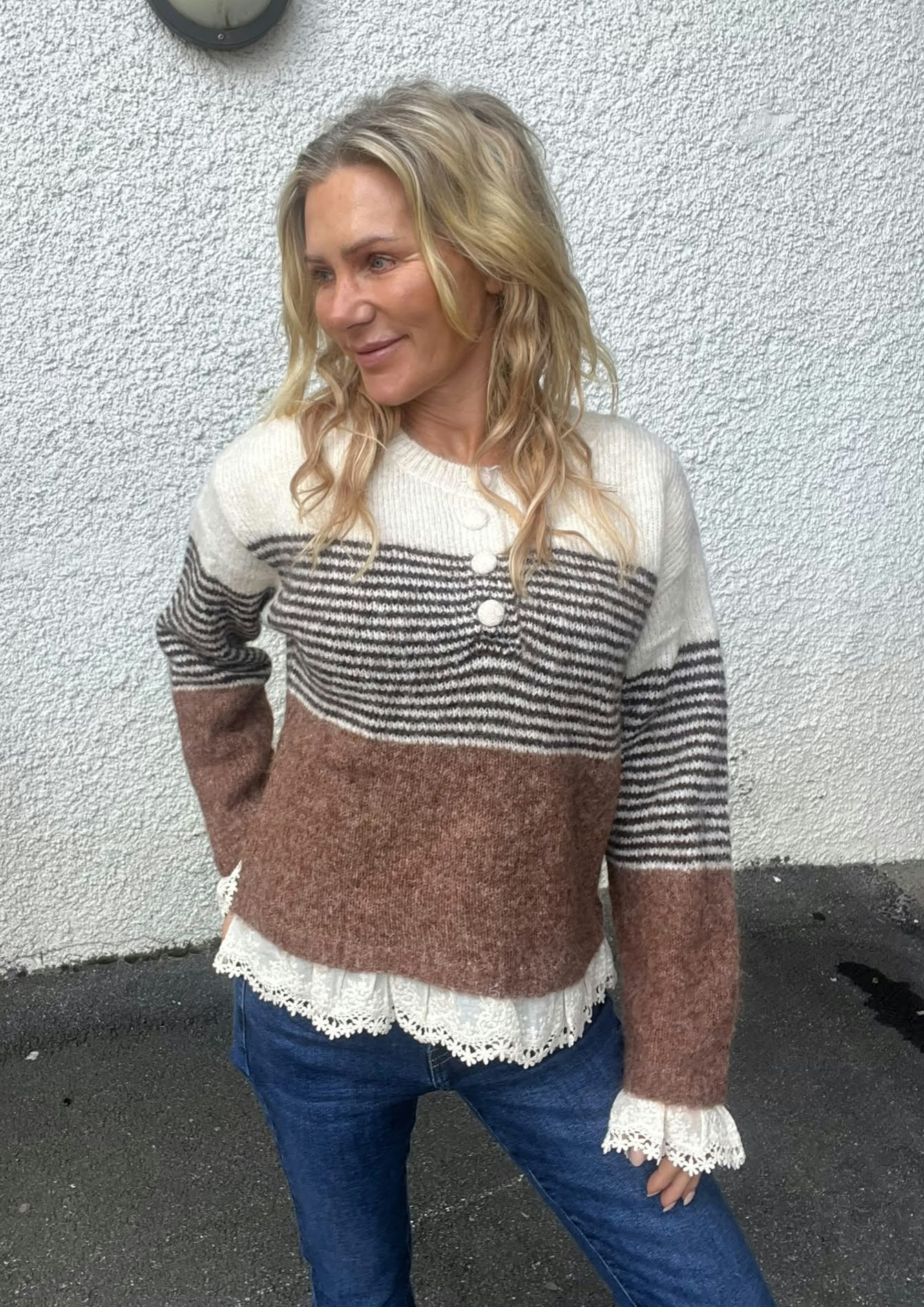 Blond kvinna står lutad mot en vit, grovputsad vägg och tittar åt vänster med ett svagt leende. Hon bär en stickad tröja med vita, svarta och bruna ränder, spetskantad blus och jeans.