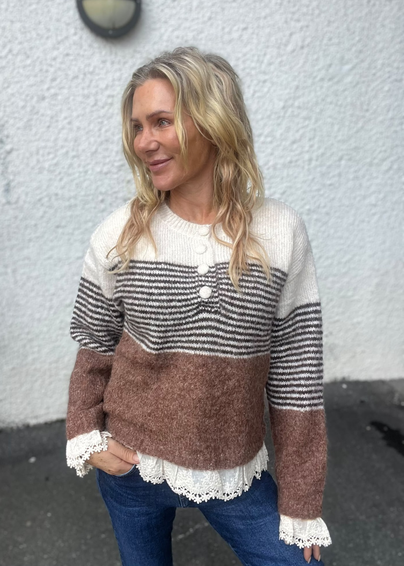 Blond kvinna står utomhus mot en vit, grovputsad vägg och ler medan hon tittar åt vänster. Hon bär en stickad tröja i vitt, grått och brunt med stora knappar, jeans och spetskantad blus under.