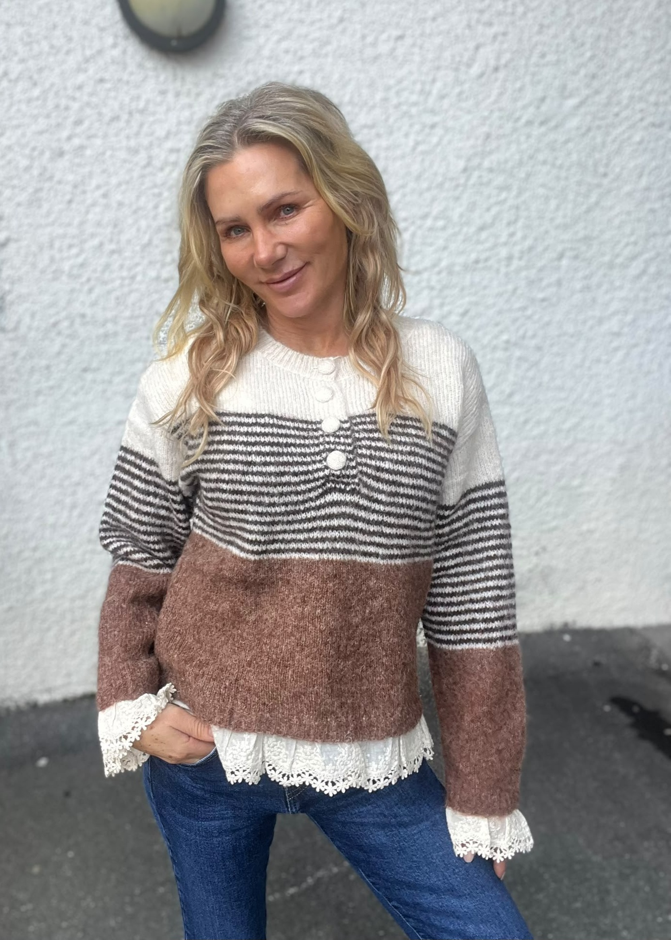 Kvinna med långt blont hår poserar och ler lätt mot kameran, med ena handen i jeansfickan. Hon står mot en vit grovputsad vägg och bär en stickad tröja med ränder och brun nederdel samt spetskant.