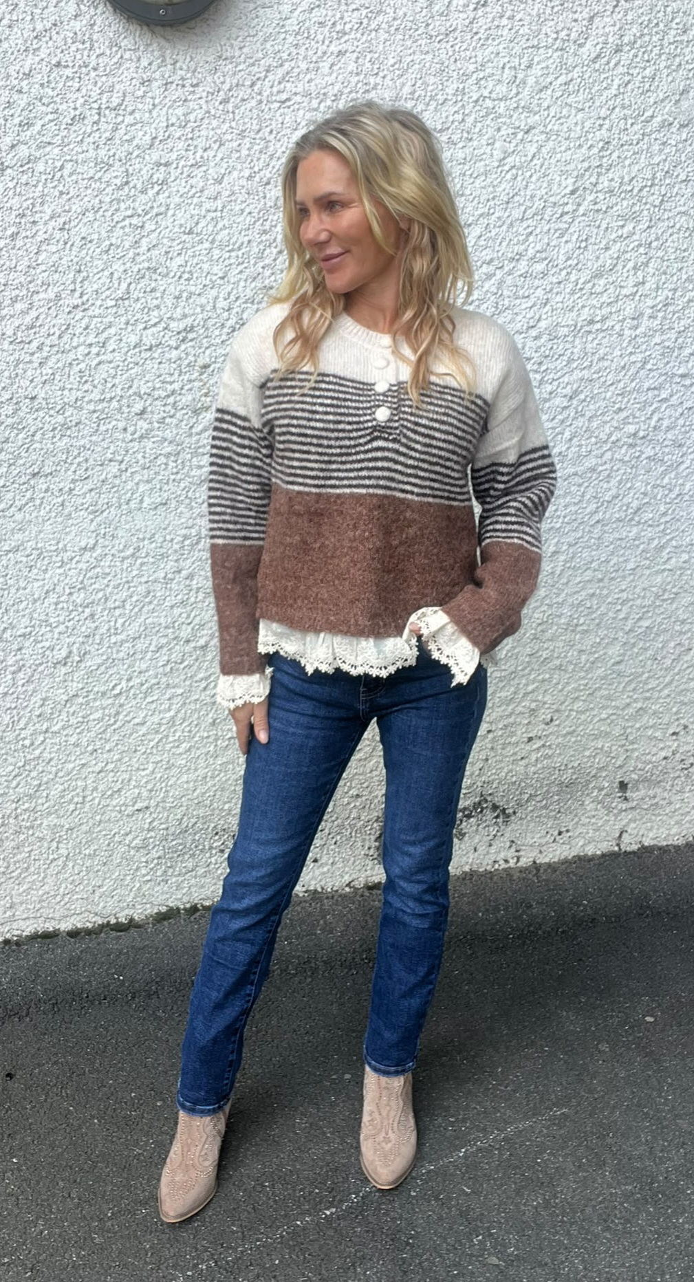 Blond kvinna står avslappnat mot en vit, grovputsad vägg och tittar åt sidan. Hon bär stickad tröja i vitt, grått och brunt med spetskant, blå jeans och ljusa boots på asfalt utomhus.