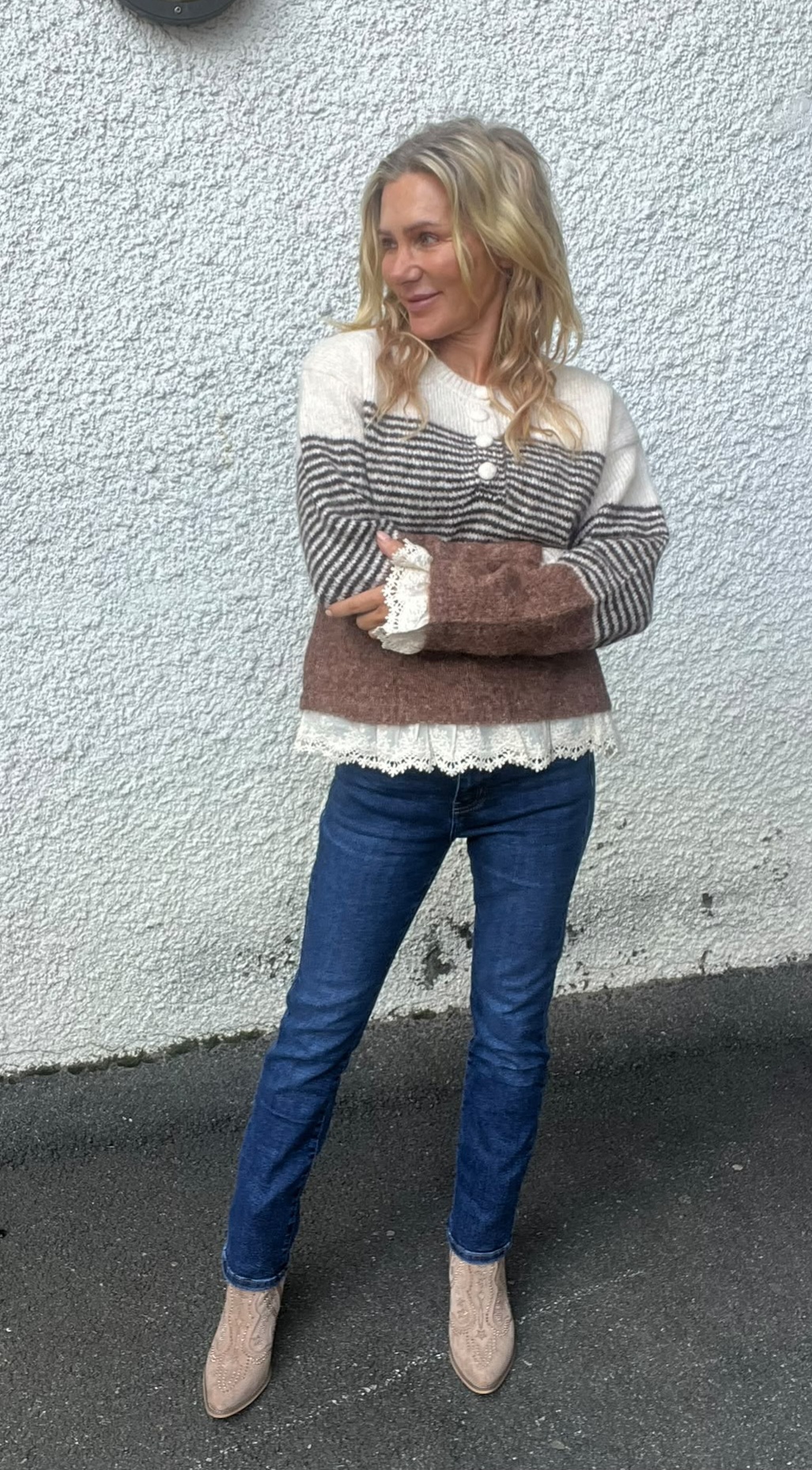 Blond kvinna står med armarna i kors och tittar åt sidan mot vänster. Hon bär randig stickad tröja med knappar och spetskant, blå jeans och ljusa boots, framför en grovt putsad ljus vägg utomhus.