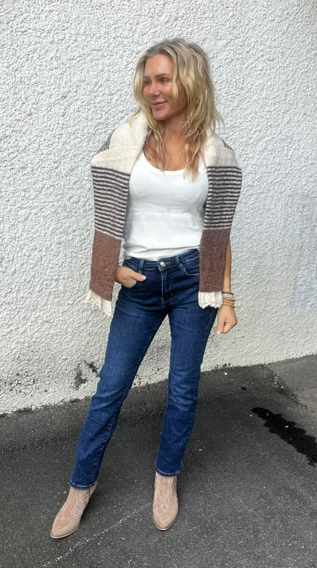 Blond kvinna står avslappnat med handen i jeansfickan och tittar åt sidan, klädd i vit topp, blå jeans och beige boots. En randig sjal eller kofta är draperad över axlarna mot en ljus, grov vägg utomhus.
