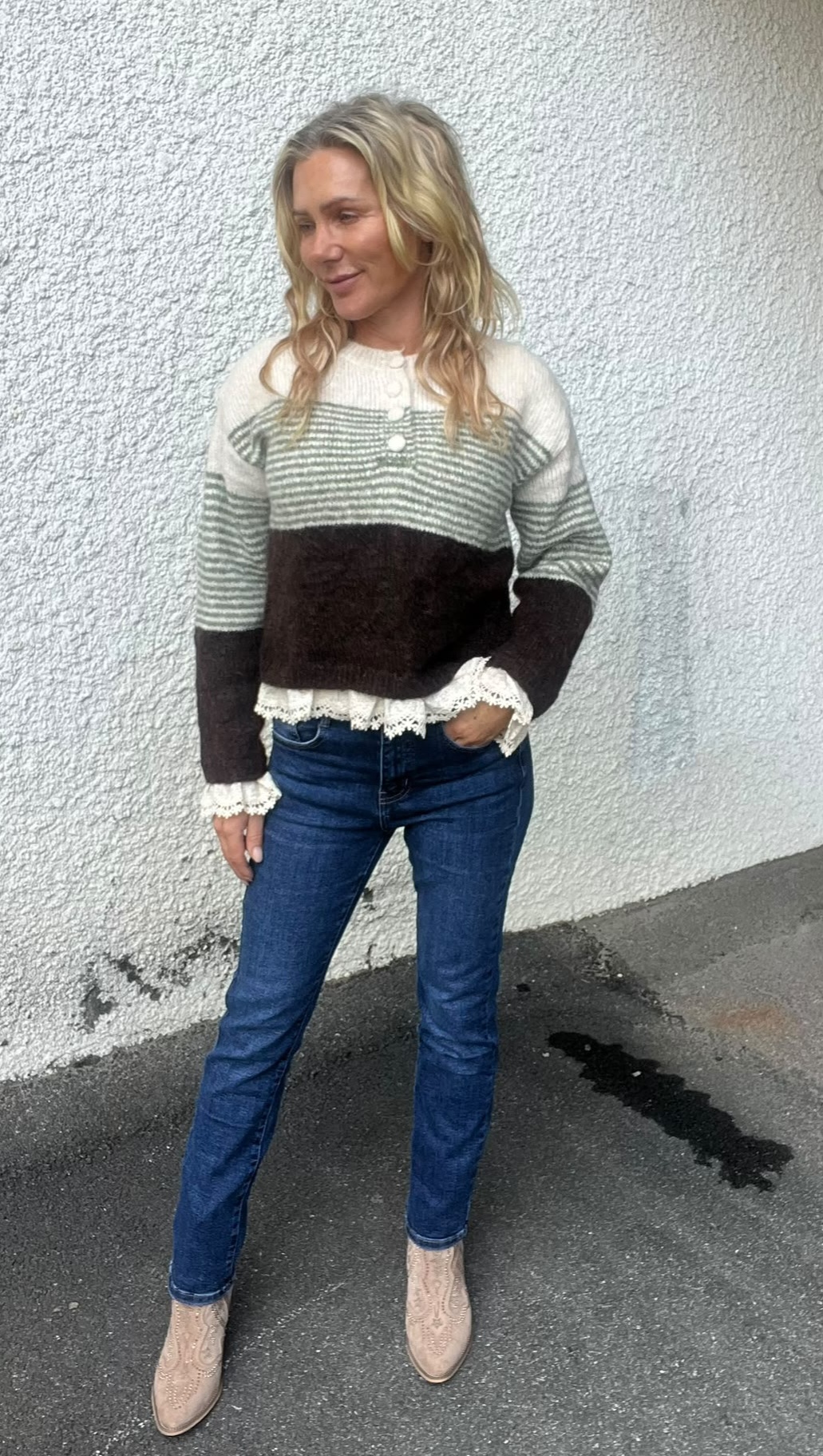 Kvinna med blont, vågigt hår står avslappnat med ena handen i fickan och tittar åt sidan, framför en vit grovputsvägg. Hon bär randig stickad tröja med spetskant, blå jeans och ljusa boots på asfalt.