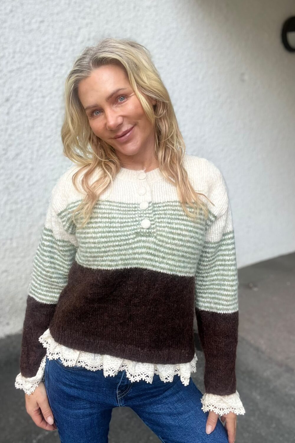 Leende kvinna med blont, vågigt hår lutar huvudet och tittar mot kameran. Hon står utomhus framför en ljus putsad vägg och bär en stickad tröja i grönt, vitt och brunt samt jeans.