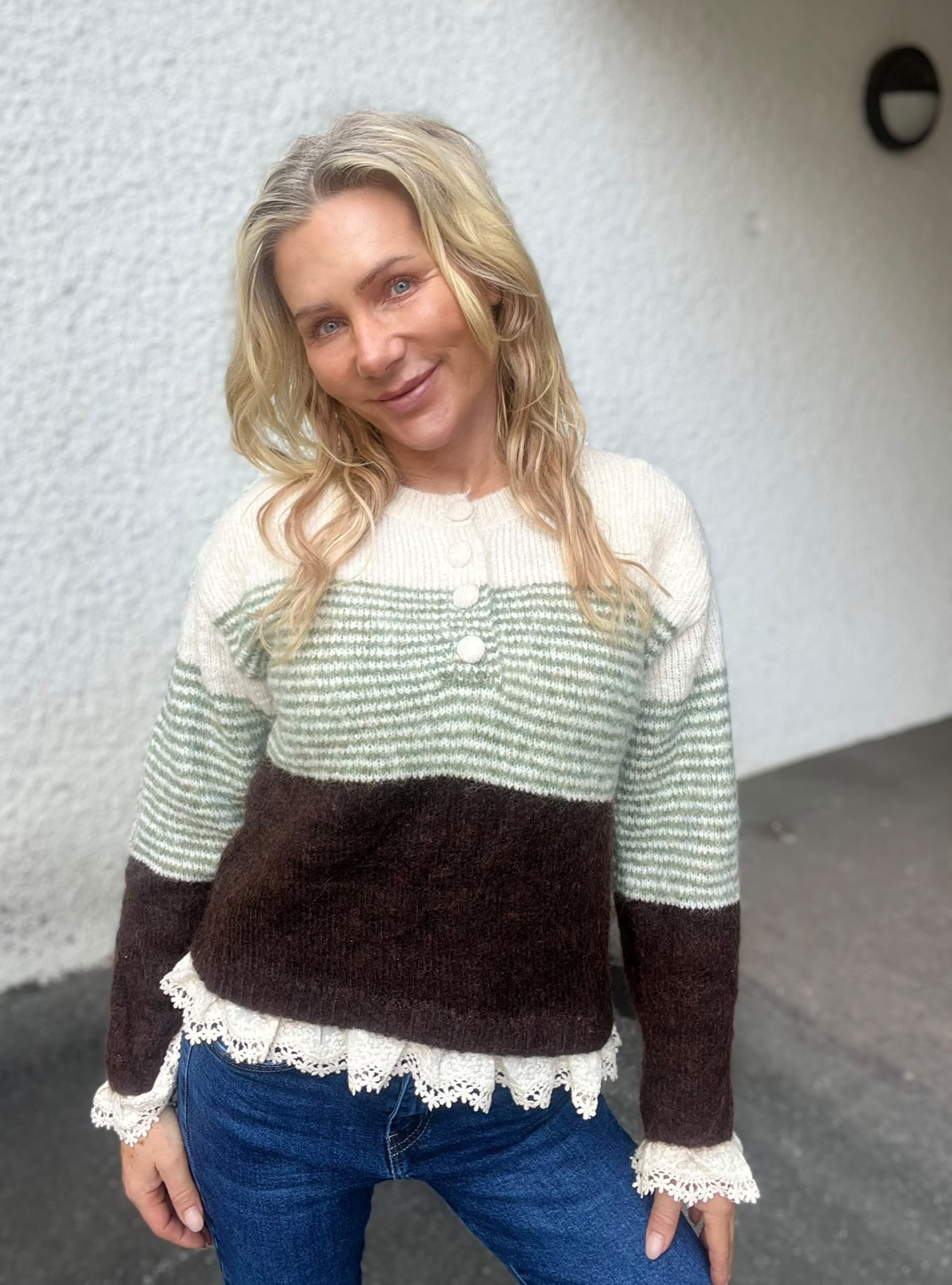 Leende kvinna med blont, vågigt hår lutar huvudet och tittar mot kameran. Hon står utomhus framför en ljus putsad vägg och bär en stickad tröja i grönt, vitt och brunt samt jeans.