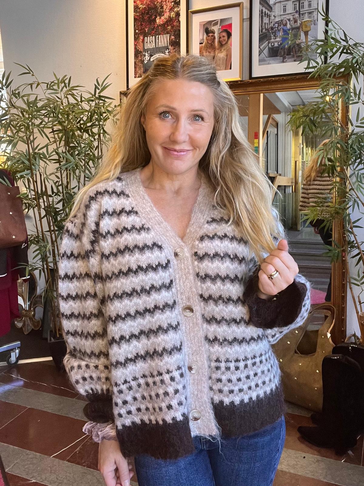 Leende kvinna med långt blont hår poserar inomhus och håller upp handen som visar en ring. Hon bär en stickad kofta med gråsvart mönster och jeans; bakom syns växter, spegel, väskor och tavlor med texten ”CASA FANNY”.