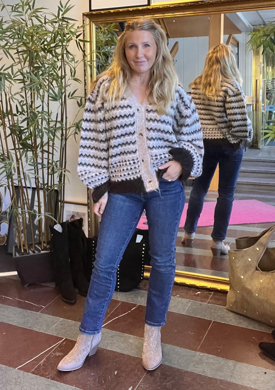 Kvinna med långt blont hår poserar framför en stor guldramsspegel, med handen i jeansfickan. Hon bär gråvit mönstrad kofta och glittriga ljusa stövletter; spegeln visar henne bakifrån i en hall med växt.