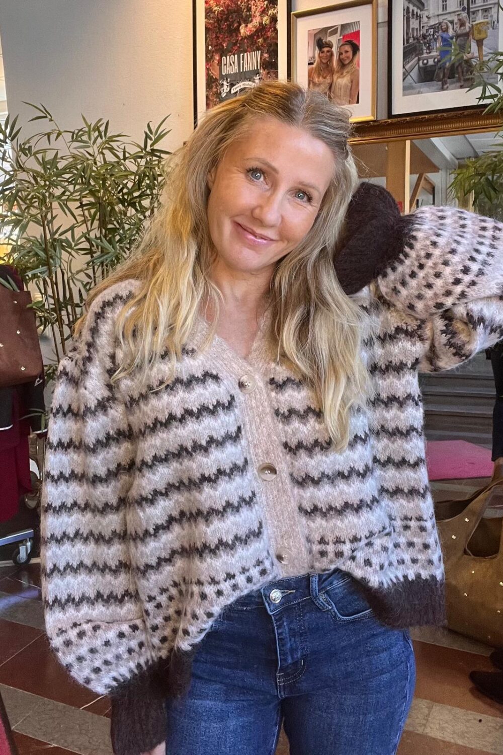 Leende kvinna med långt blont hår poserar med ena armen bakom huvudet och ser in i kameran, iklädd mönstrad stickad kofta och jeans, inomhus bland växter och inramade foton; texten “CASA FANNY” syns.
