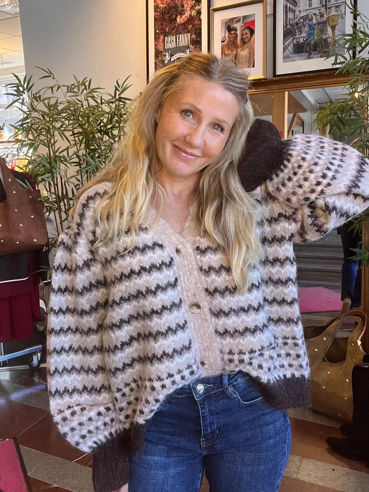 Leende kvinna med långt blont hår poserar med ena armen bakom huvudet och ser in i kameran, iklädd mönstrad stickad kofta och jeans, inomhus bland växter och inramade foton; texten “CASA FANNY” syns.